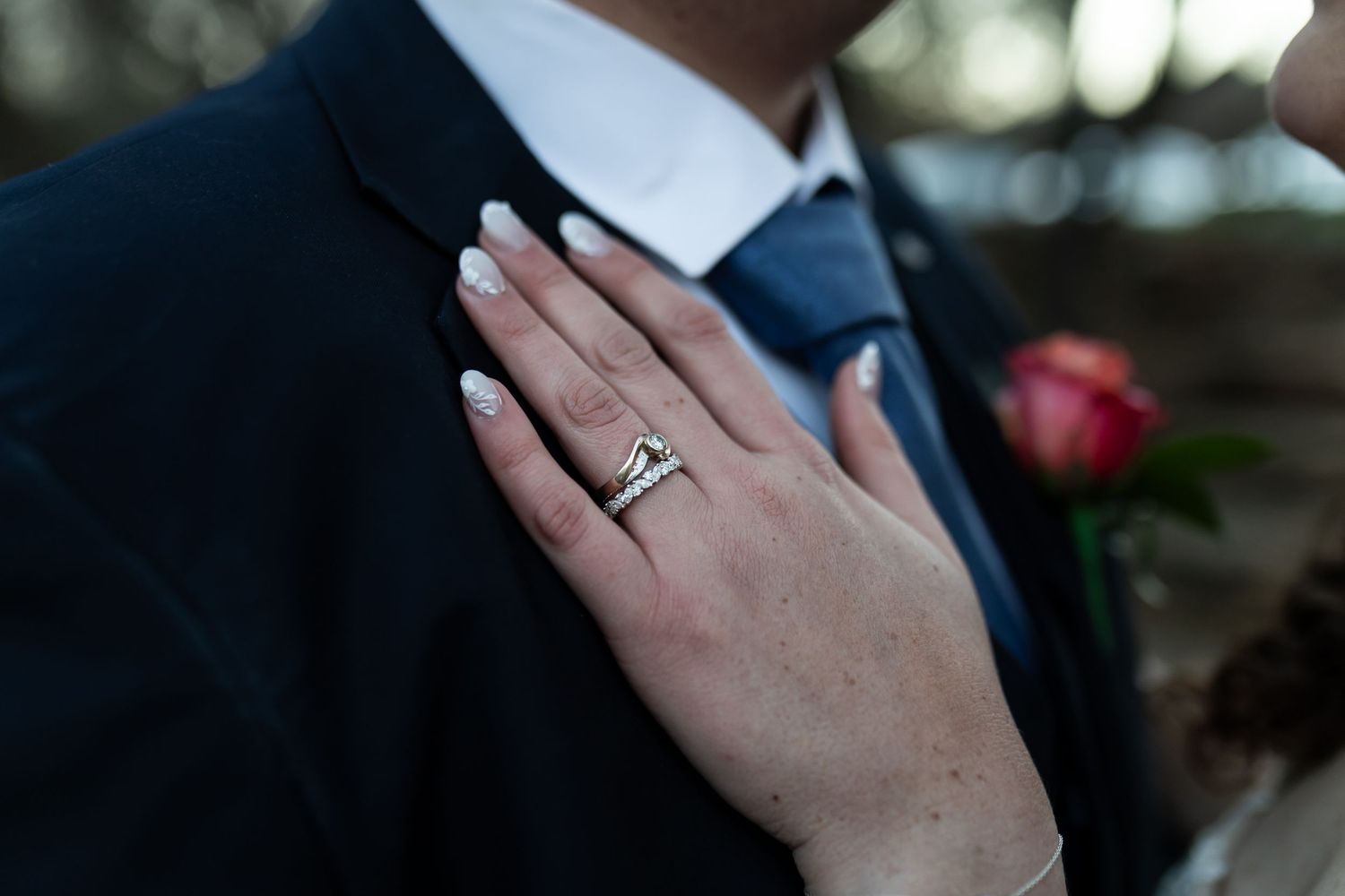 A feminine hand with an engagement ring rests on a dark suit lapel next to a red rose.
