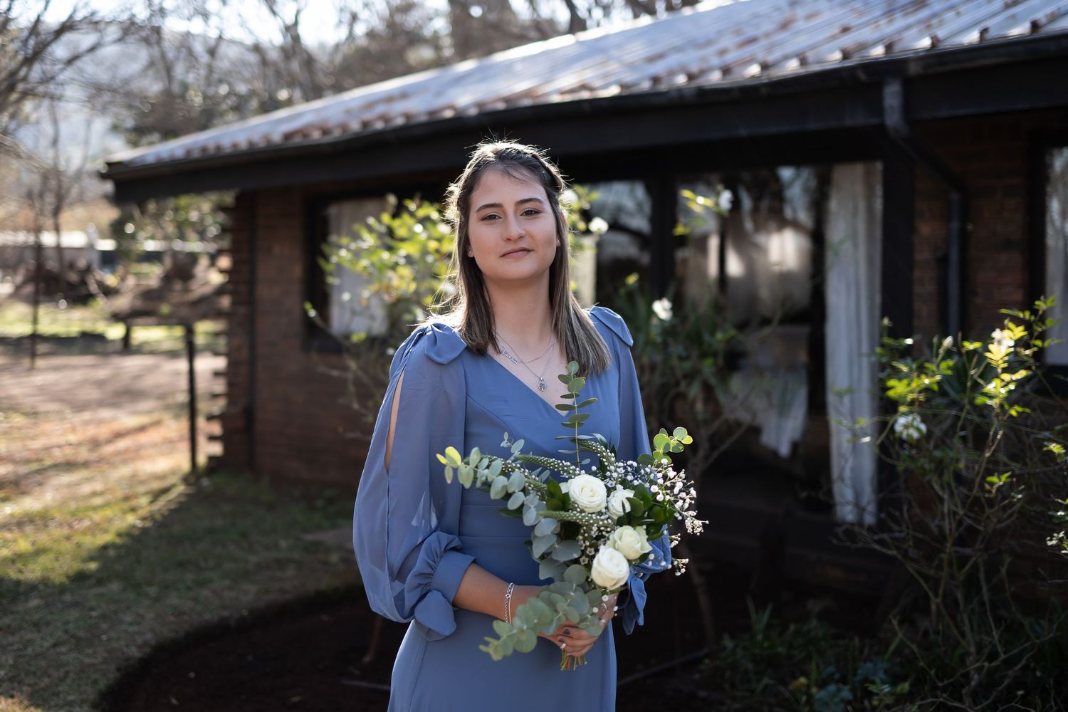 Someone in a blue dress holds a floral bouquet while standing outside a rustic building on a sunny day.