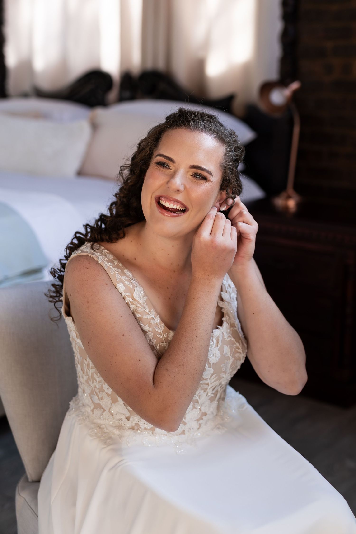 A bride in a lace wedding gown laughs and smiles while sitting in an elegant indoor setting.