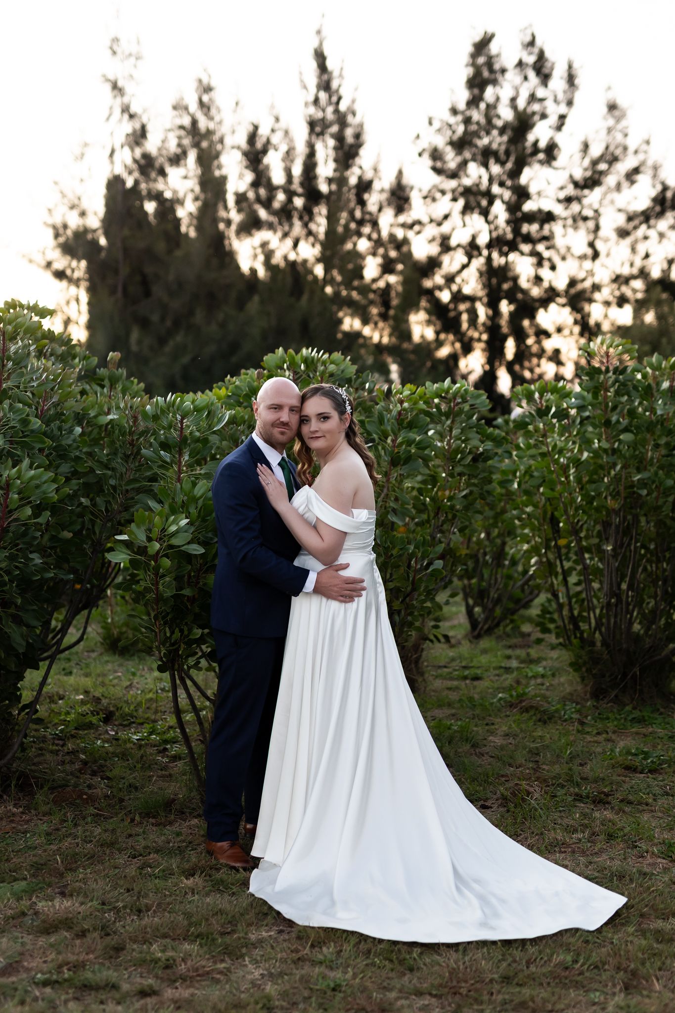 An elegant full-length wedding portrait against a backdrop of garden greenery at sunset.