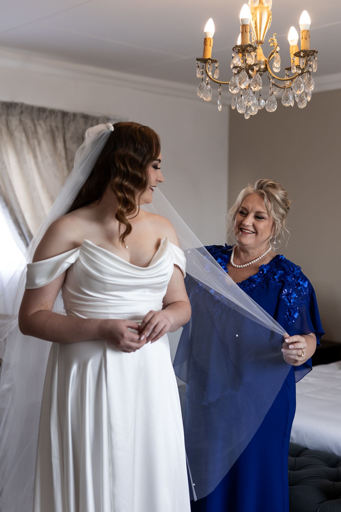 Elegant chandelier illuminates wedding preparation with helper adjusting long tulle veil.