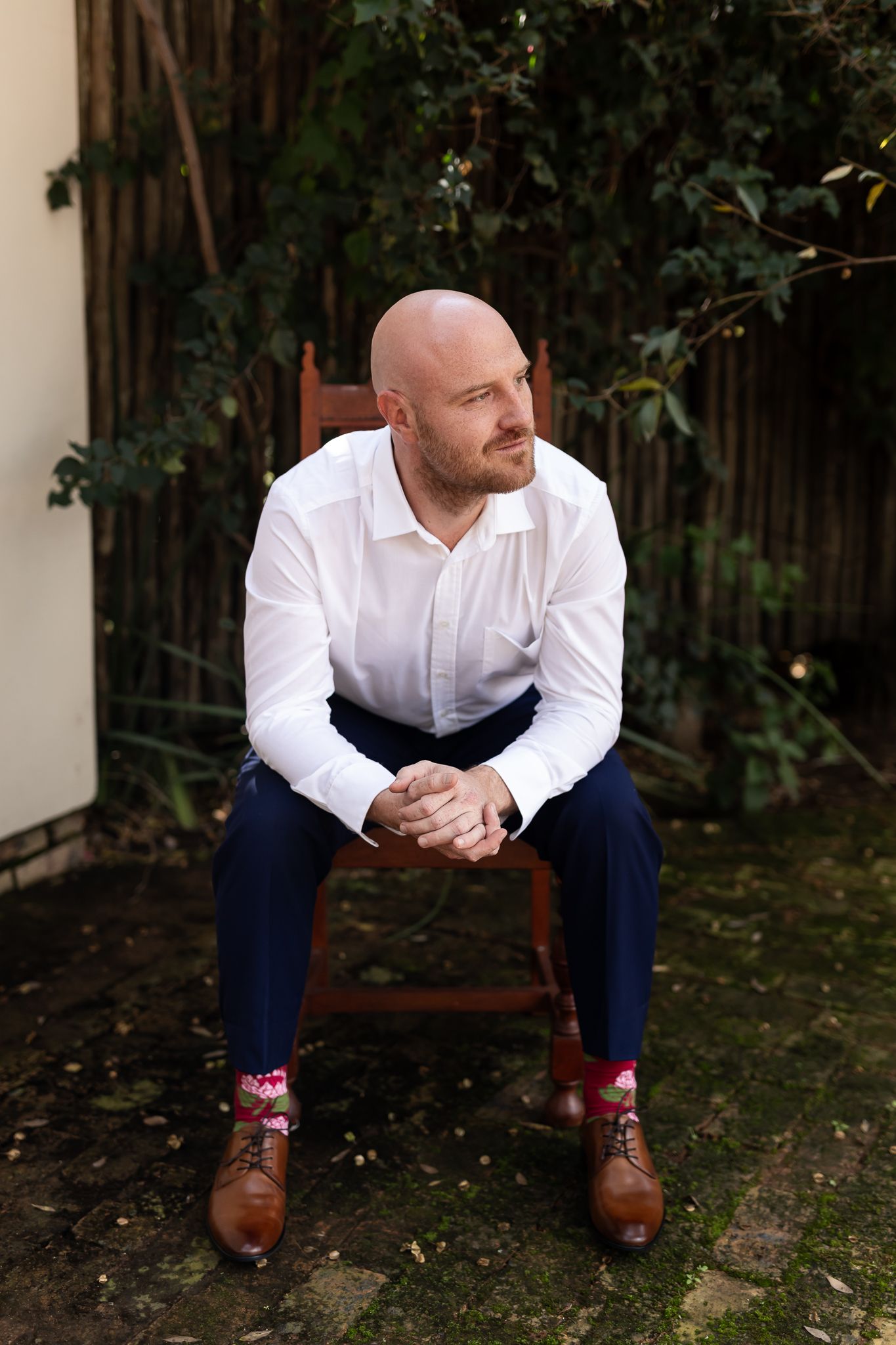 Groom in white shirt and navy pants sitting on wooden chair in garden setting.