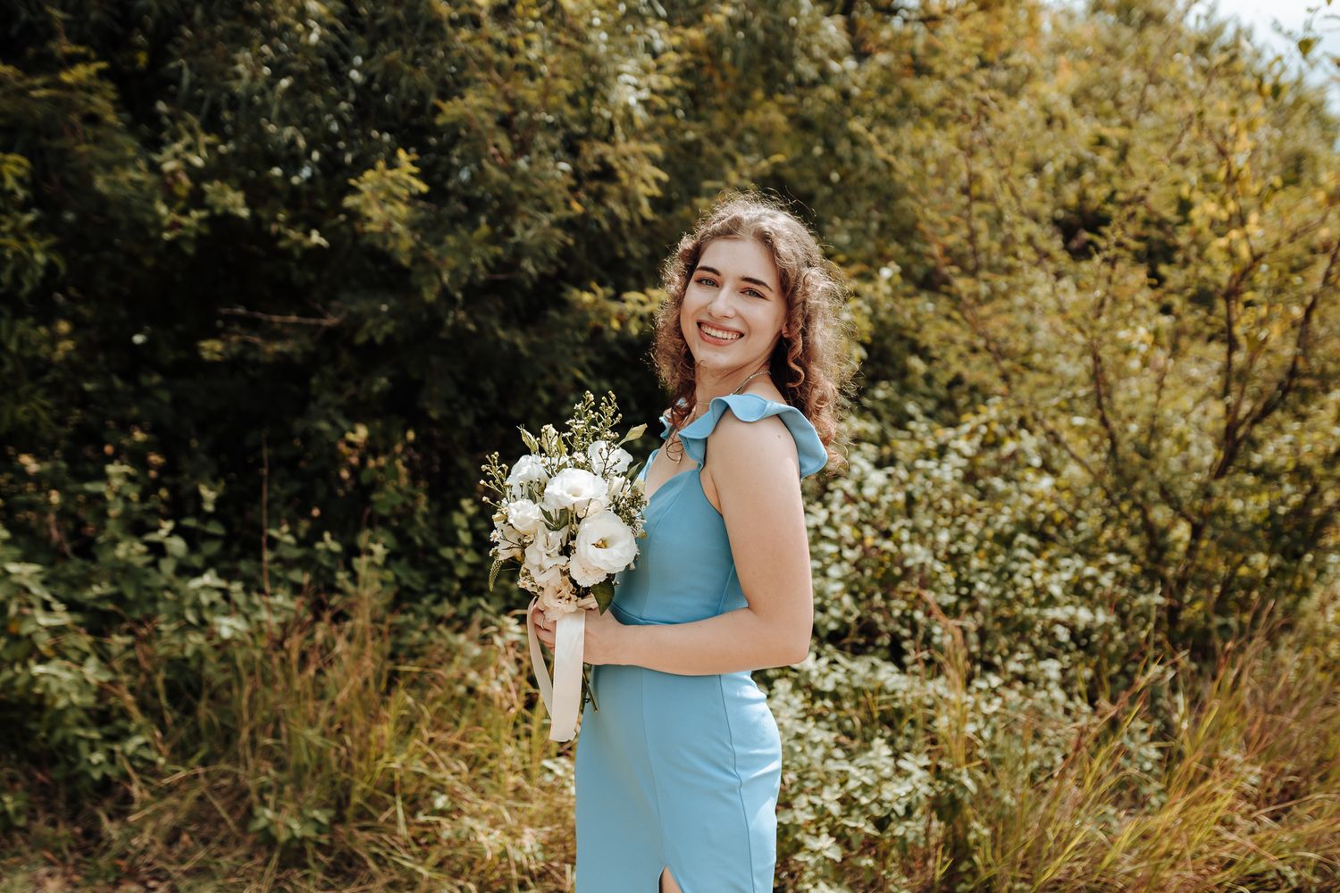 Person in light blue dress holding white floral bouquet against greenery backdrop.