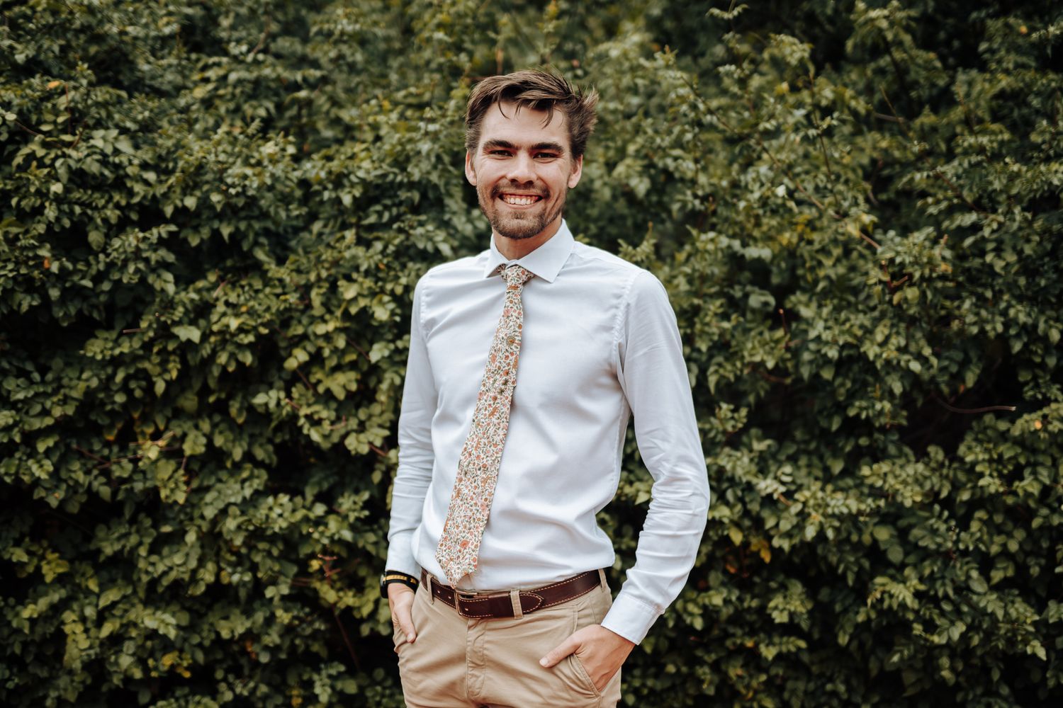 Groomsman in white button-up and floral tie smiles against leafy background.