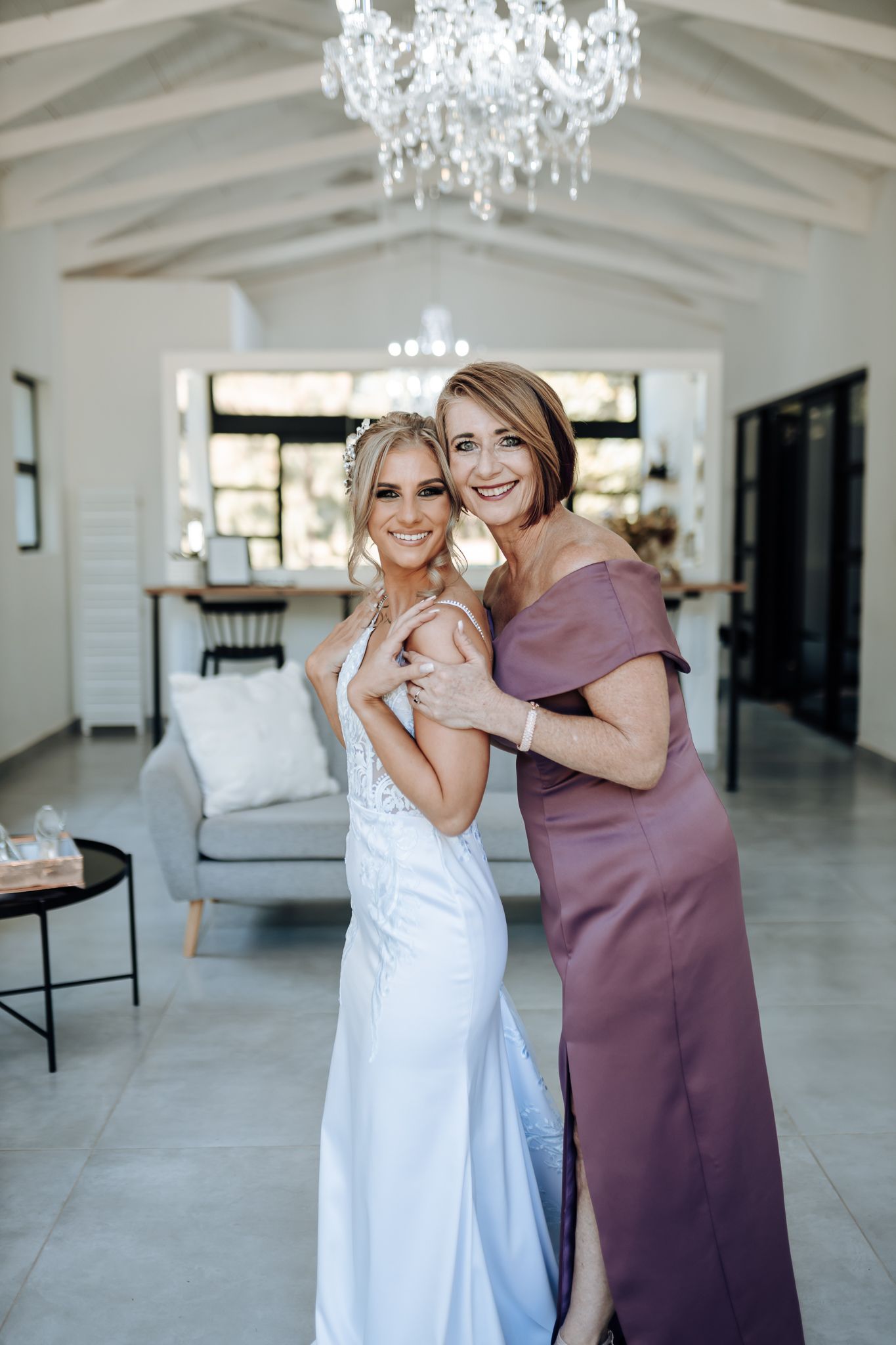 Two people share joyful moment under crystal chandelier in modern white room during wedding preparations.