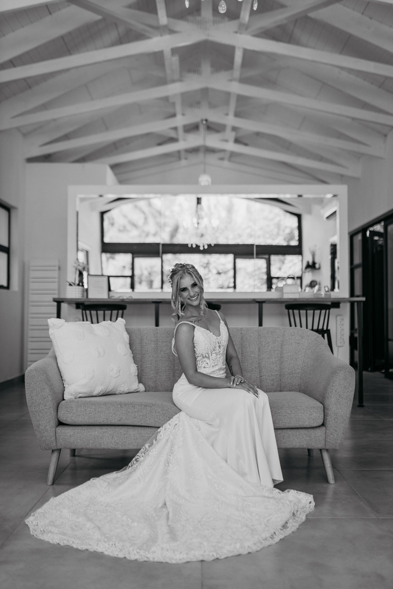 Black and white portrait of bride in lace gown seated on modern gray couch.