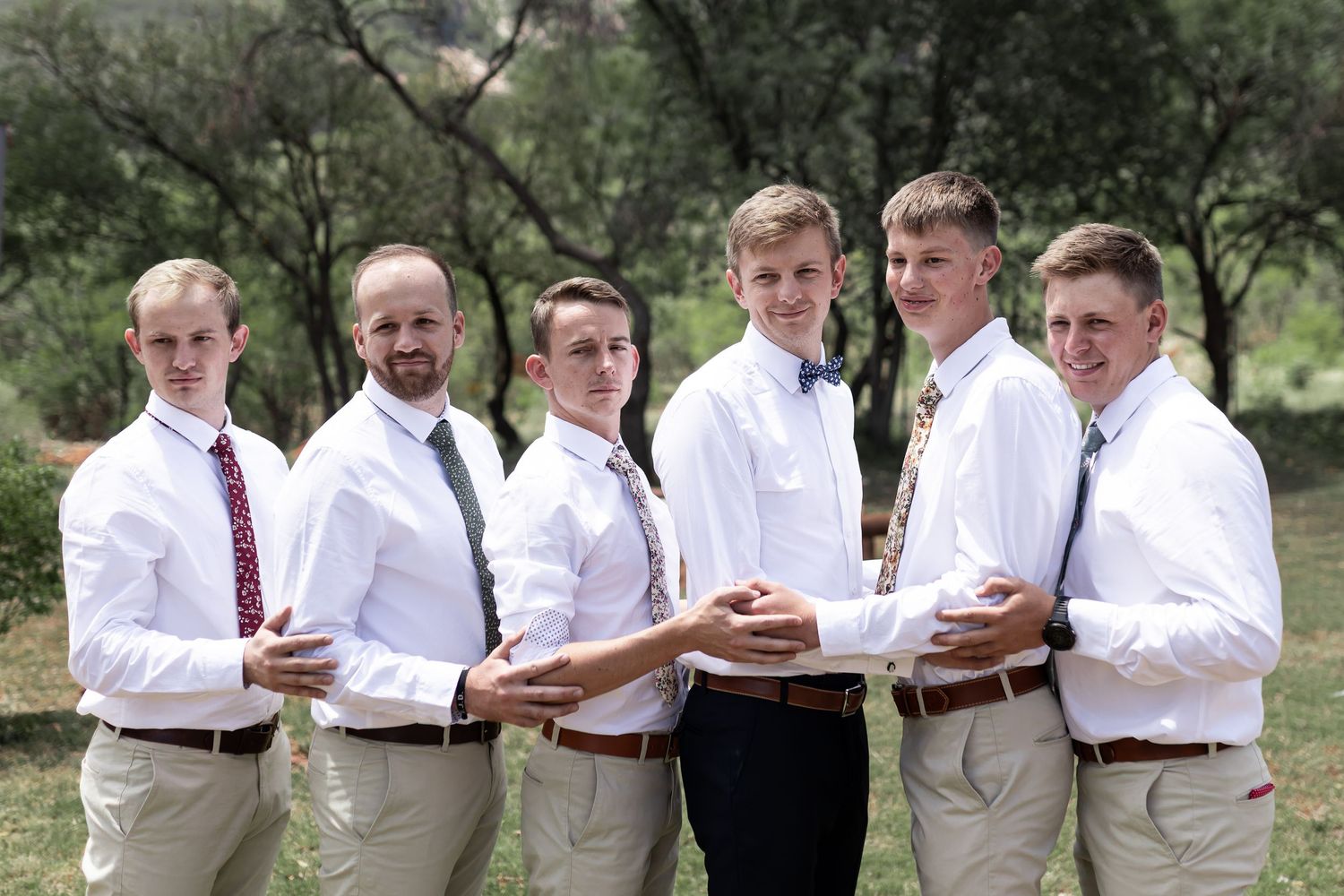 Wedding party in white shirts and khakis poses casually outdoors with drinks.