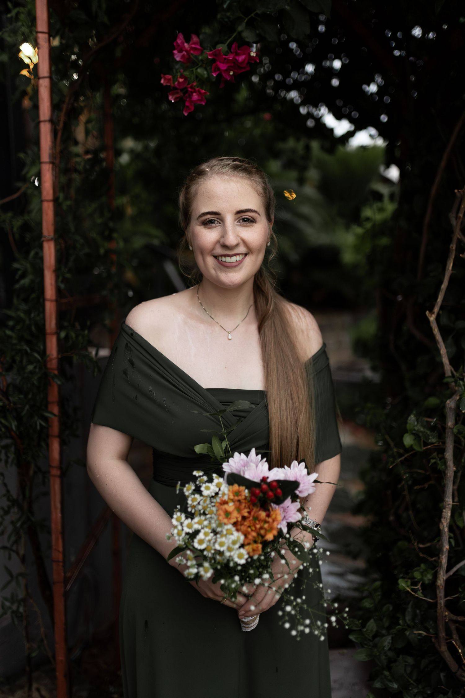 Bridesmaid in dark green dress holding colorful wildflower bouquet in garden setting.