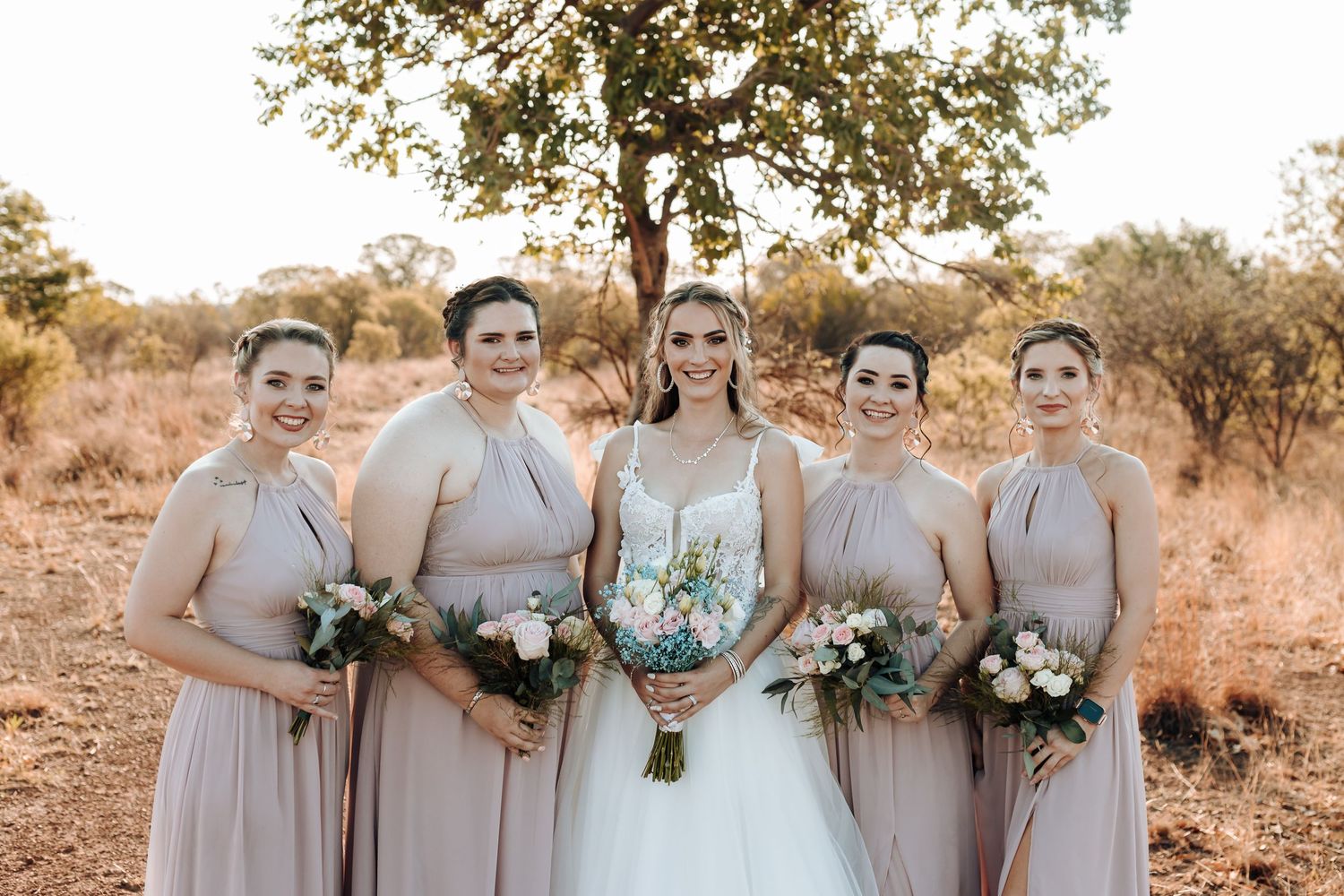 Bridal party in soft mauve dresses posing with bouquets in outdoor setting.