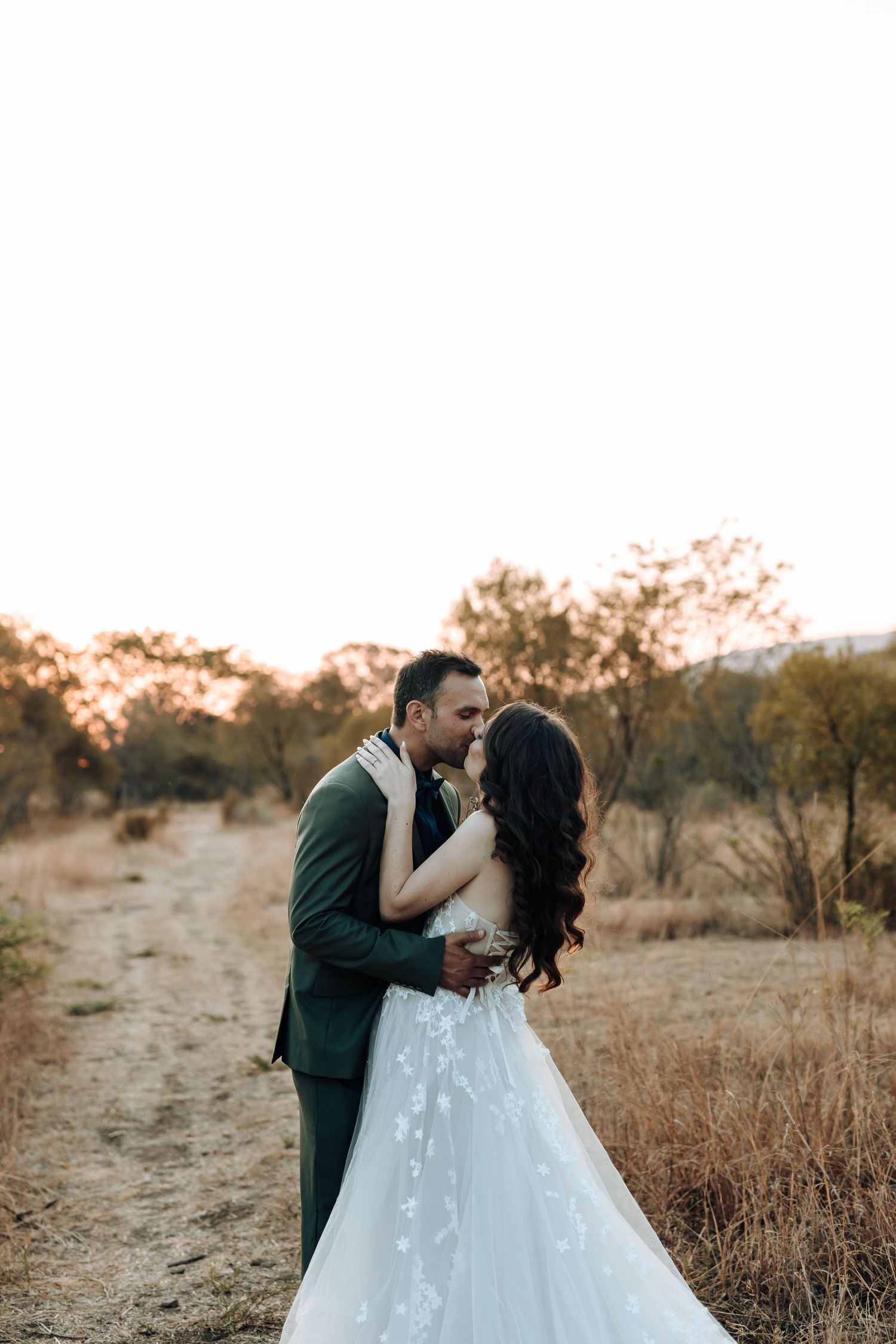 Newlyweds share an intimate moment on a dirt path at golden hour in a rustic outdoor setting.