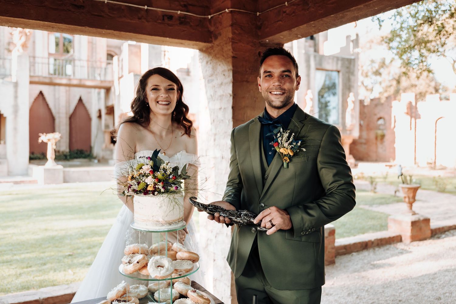 Wedding couple poses with their cake and desserts under a covered outdoor reception area.