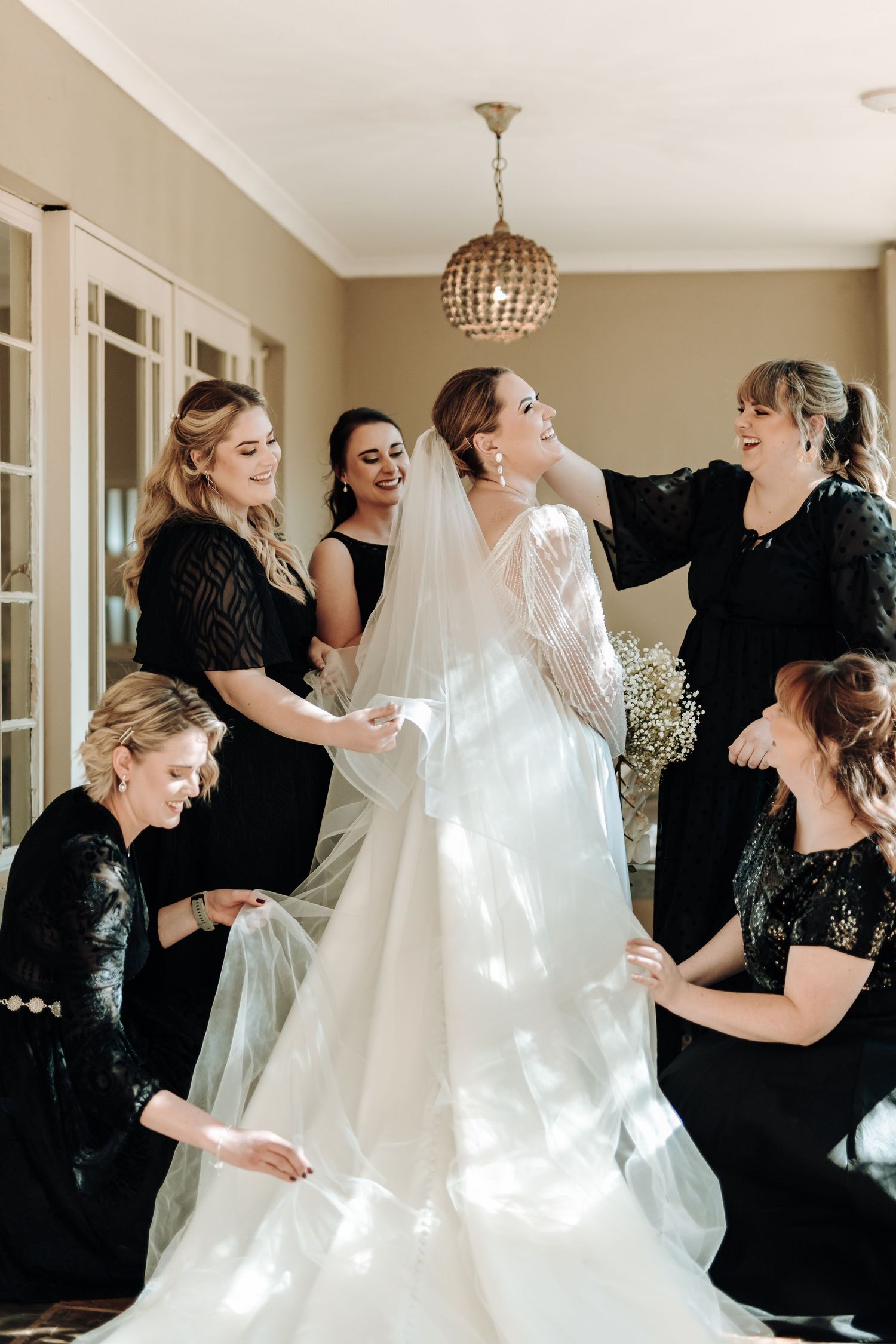 A bridal party helps adjust a white wedding dress in a sunlit room with a crystal chandelier.