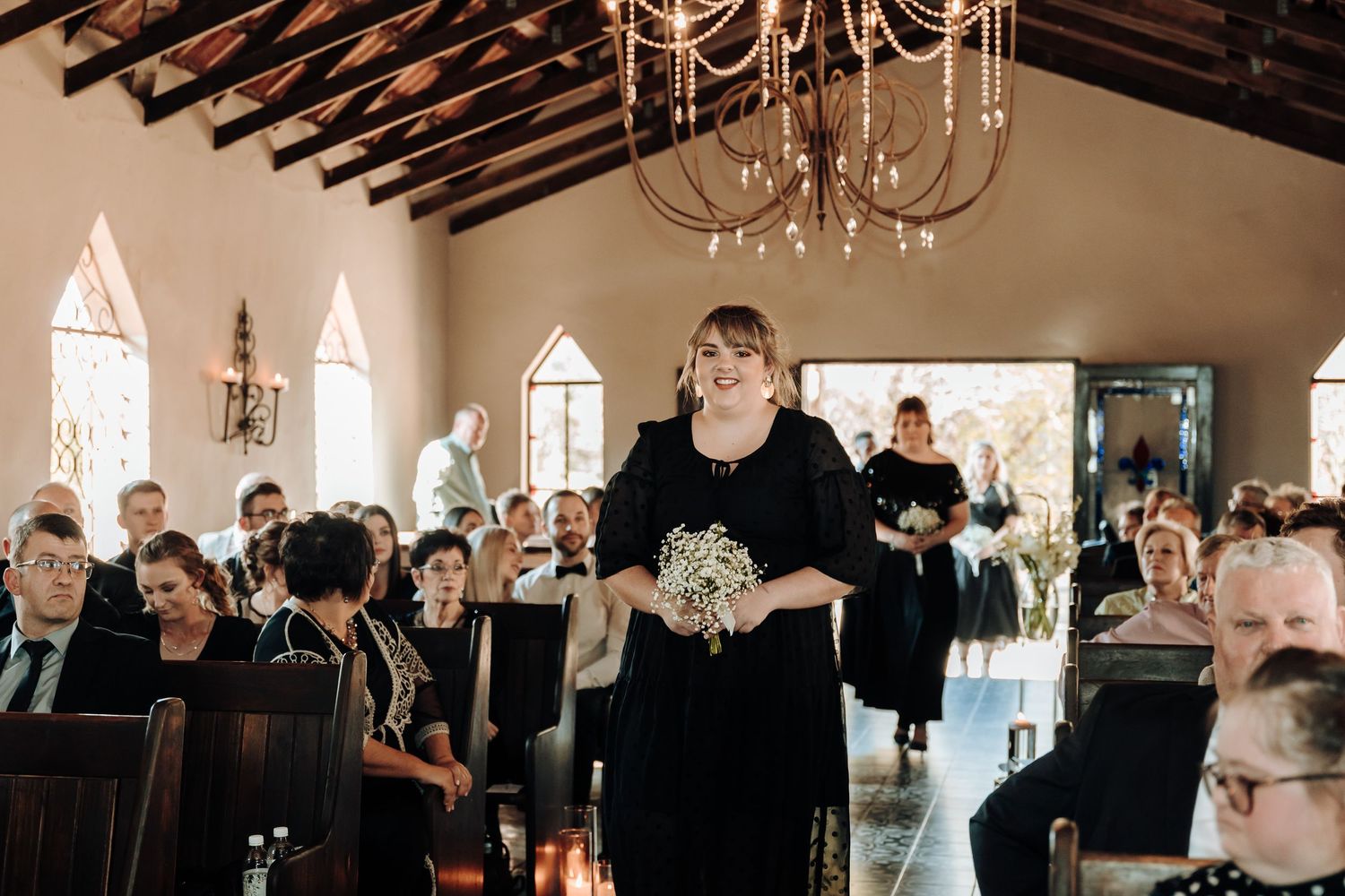 Wedding ceremony view with bridesmaids in black dresses walking down church aisle.