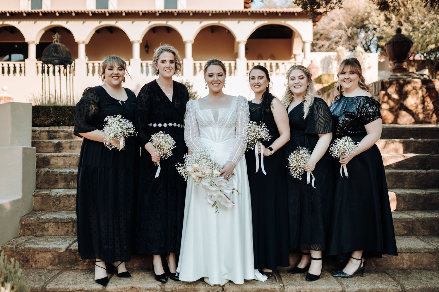Bridal party in black dresses poses formally on stone steps of an arched building.