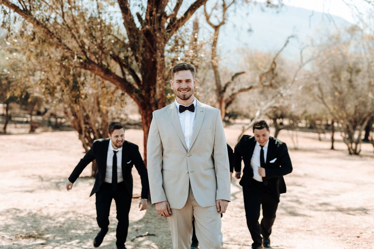 Wedding party walks behind someone in a light colored suit outdoors.