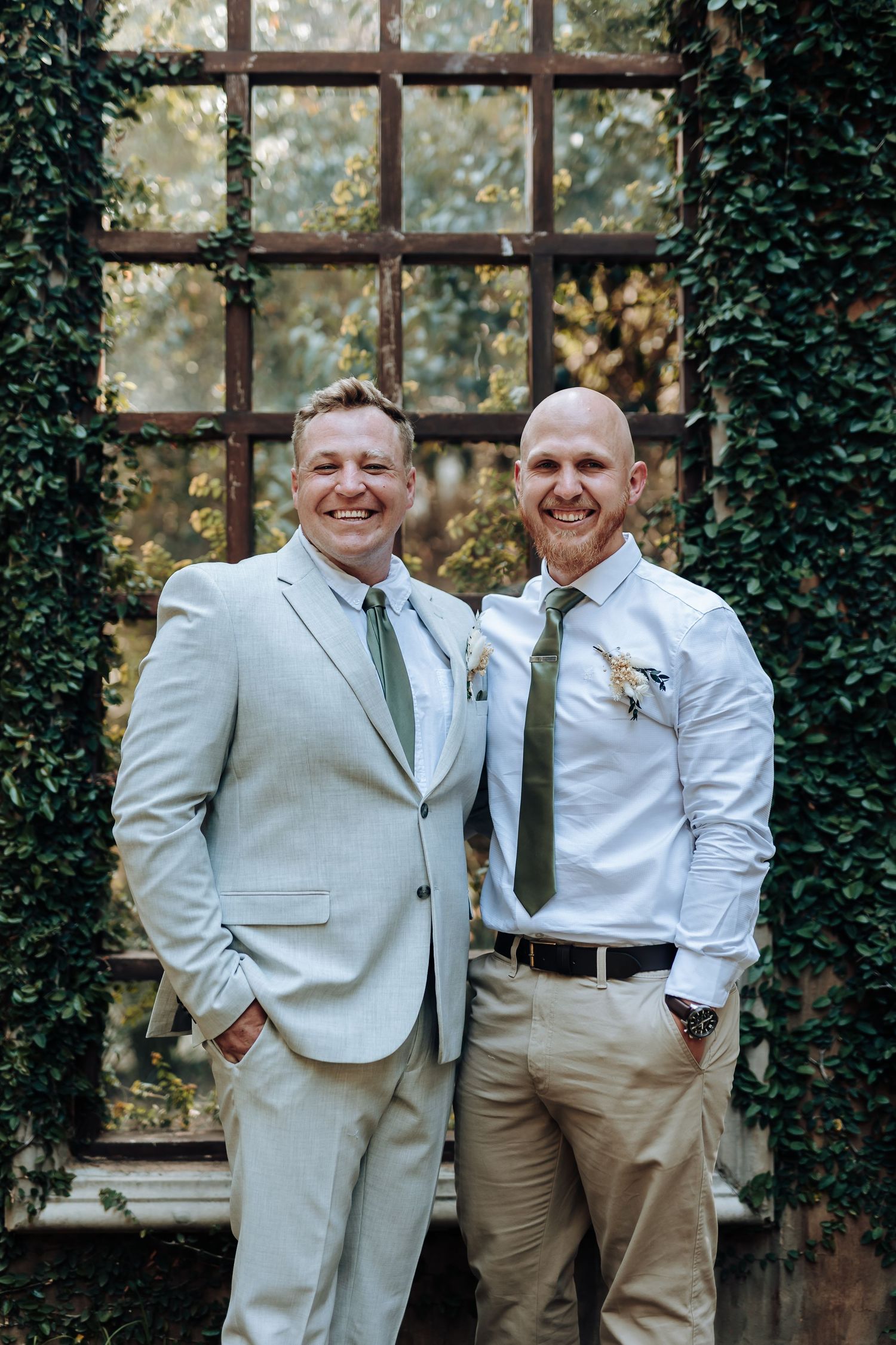 Two people in light formal attire smile while standing in front of a rustic window frame with ivy.