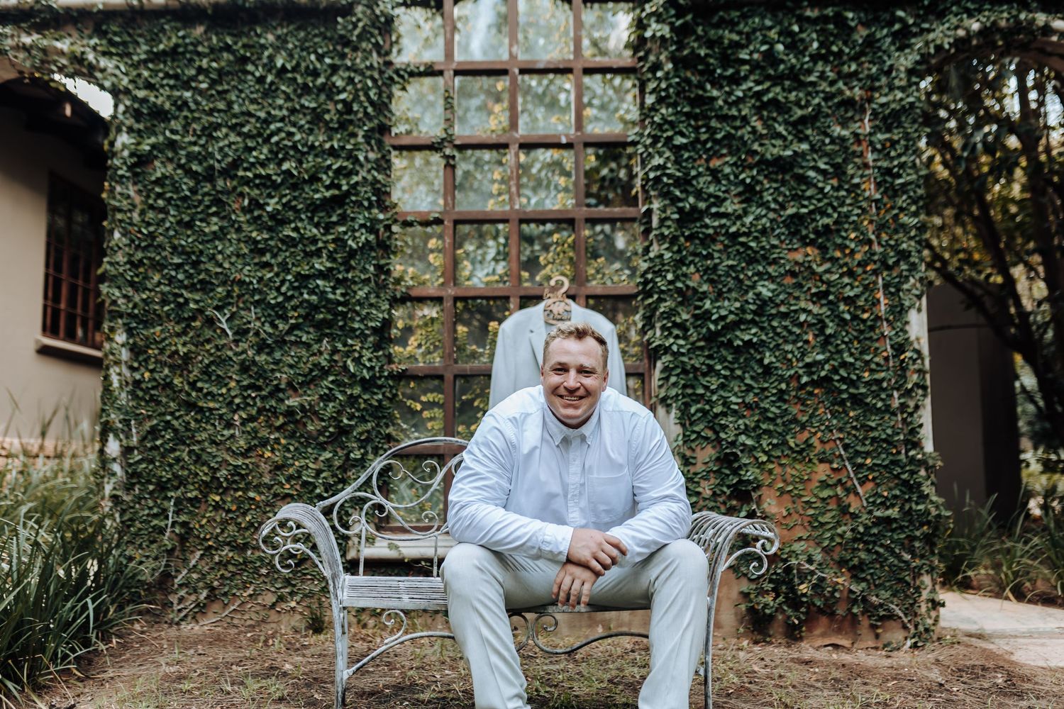 Man in light gray suit sits casually on bench against ivy-covered wall.