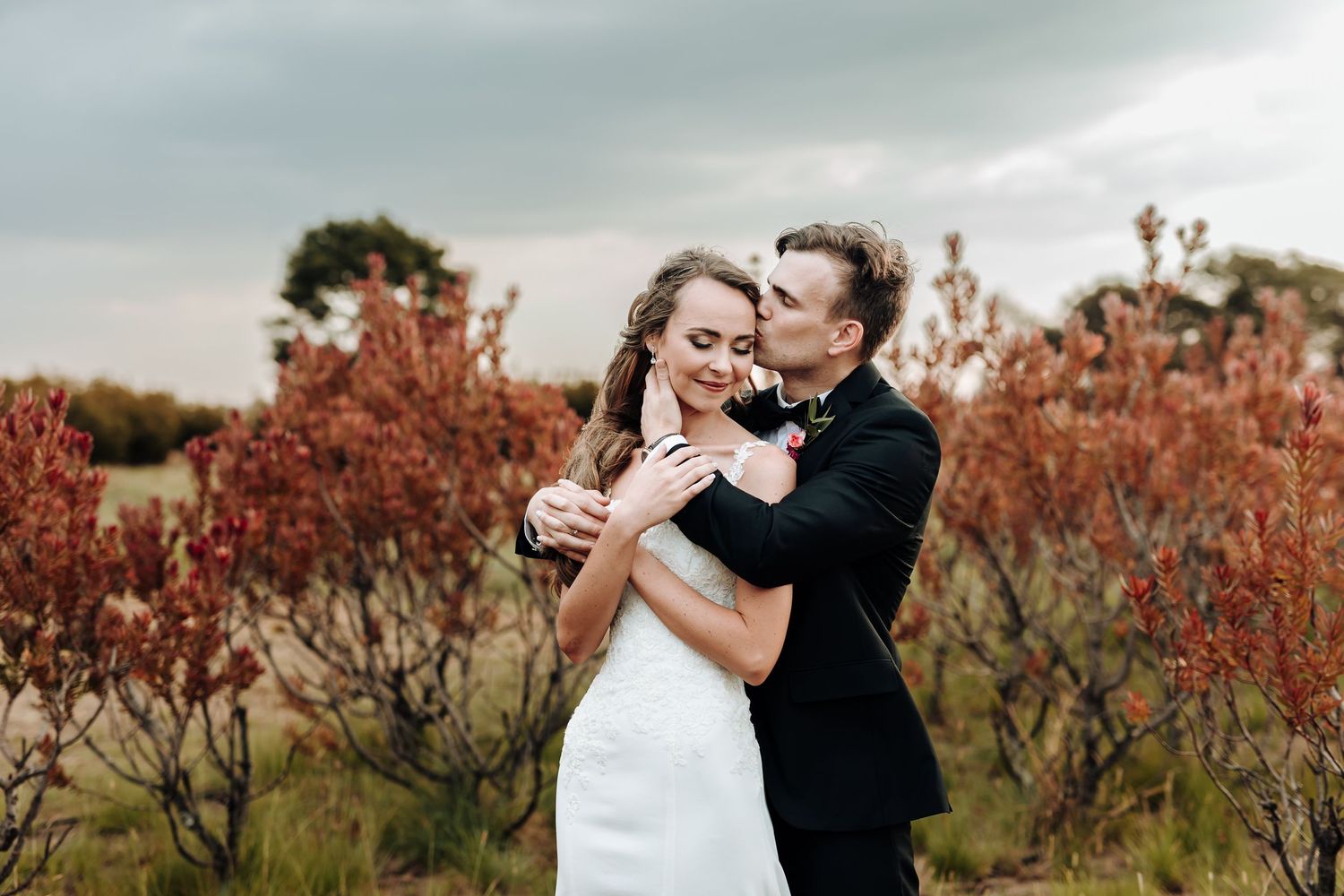 Newlyweds embrace in a field of vibrant red autumn foliage against a moody sky.