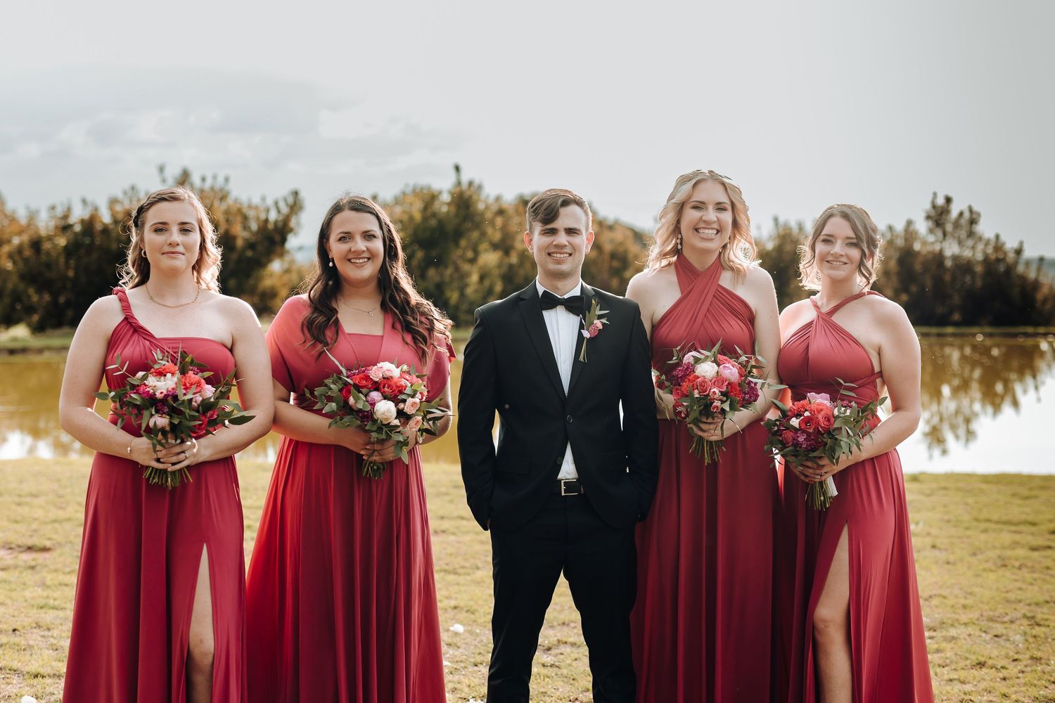 Bridesmaids in flowing burgundy gowns pose with bouquets by lakeside.