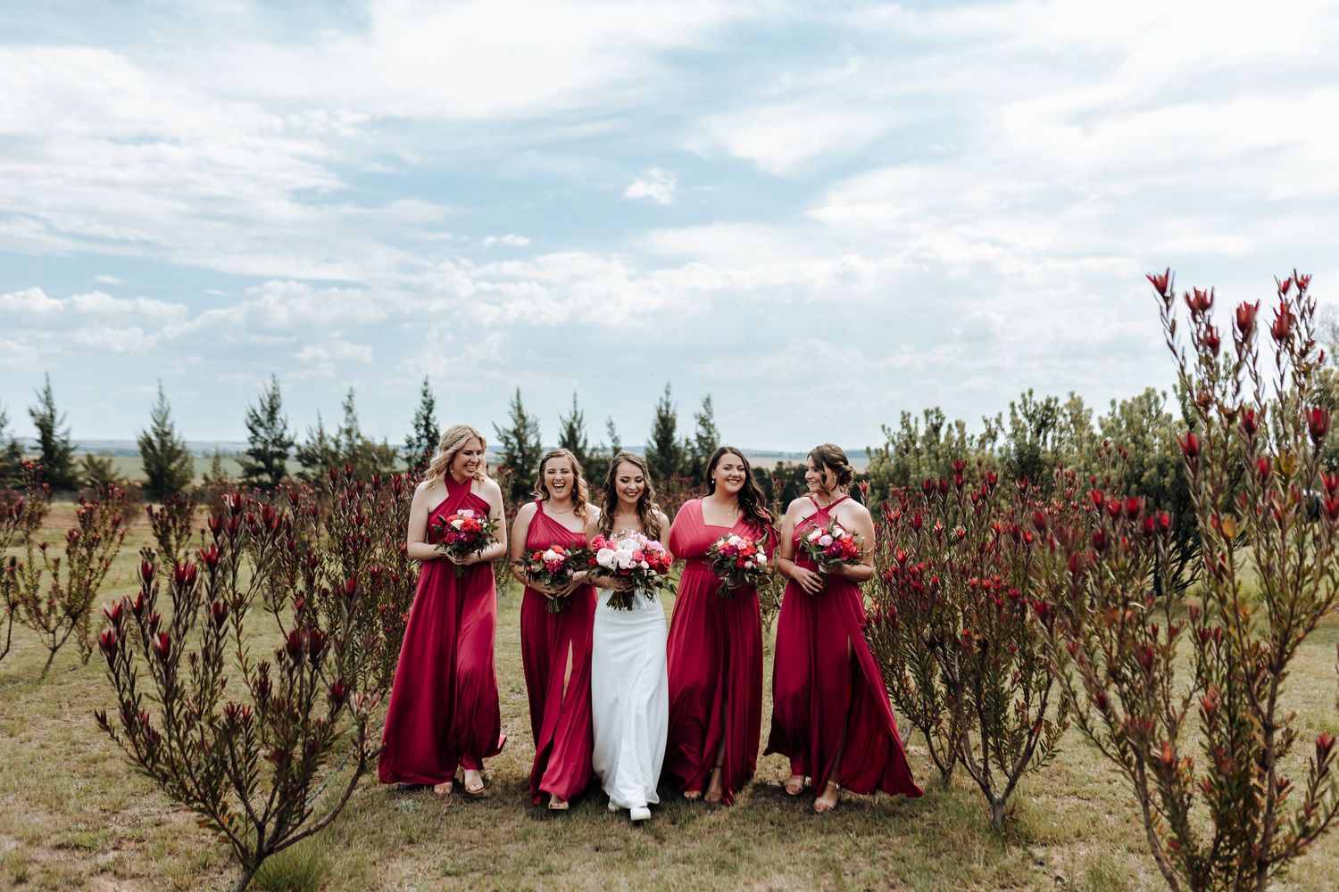 Wedding party in red dresses stands together in outdoor garden setting.