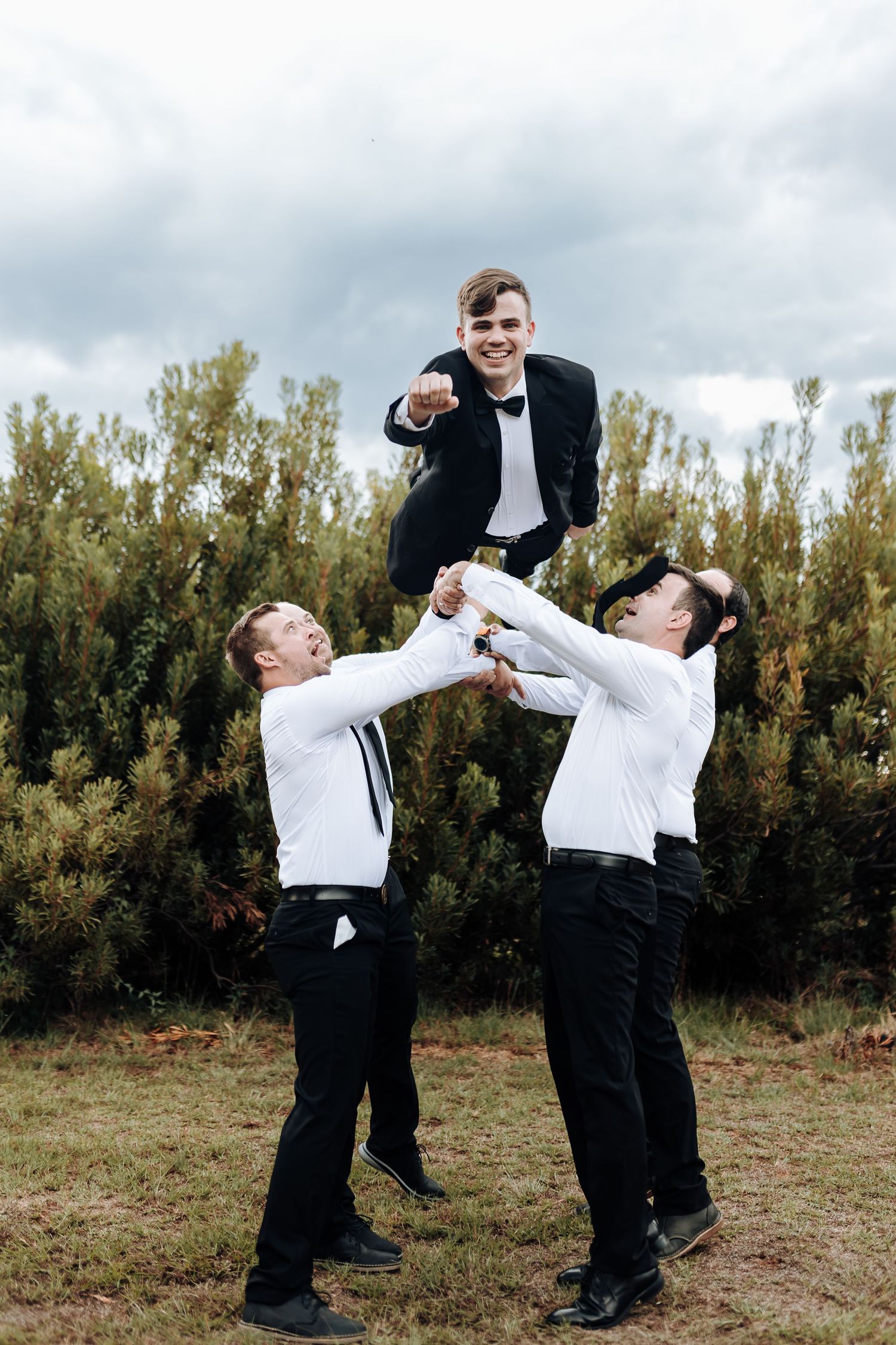 Three groomsmen perform a cheerful lifting pose outdoors during wedding photography.