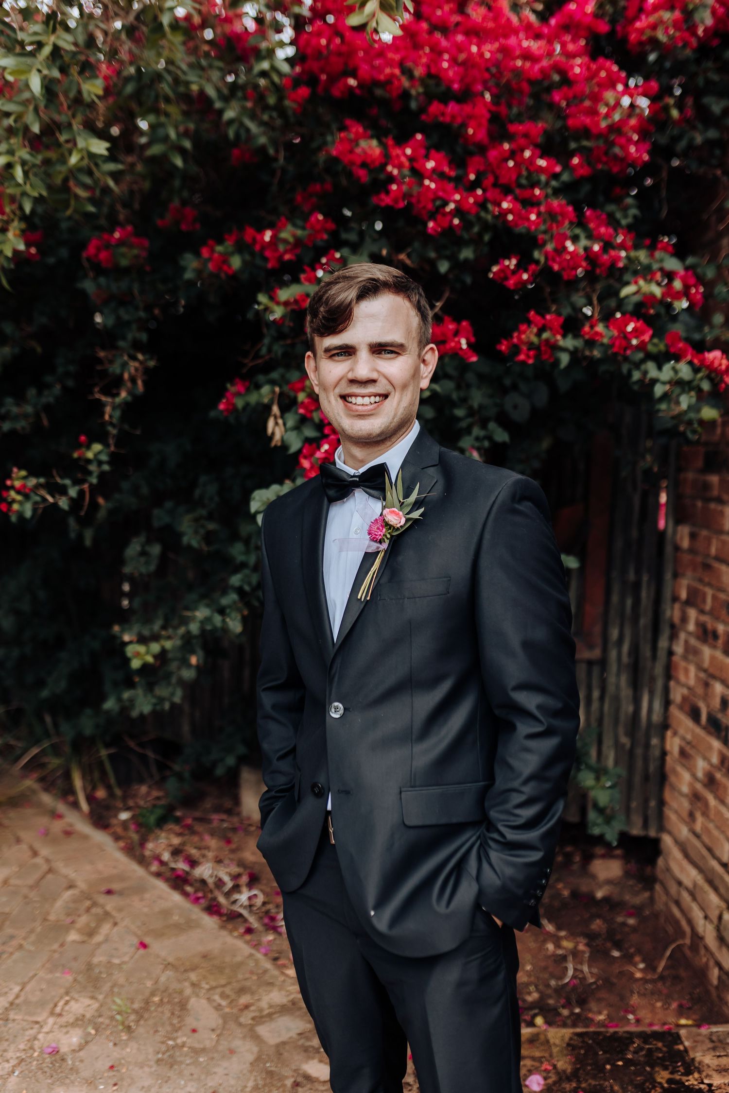 A formal black tuxedo with red boutonniere poses against vibrant red flowers and brick wall background.