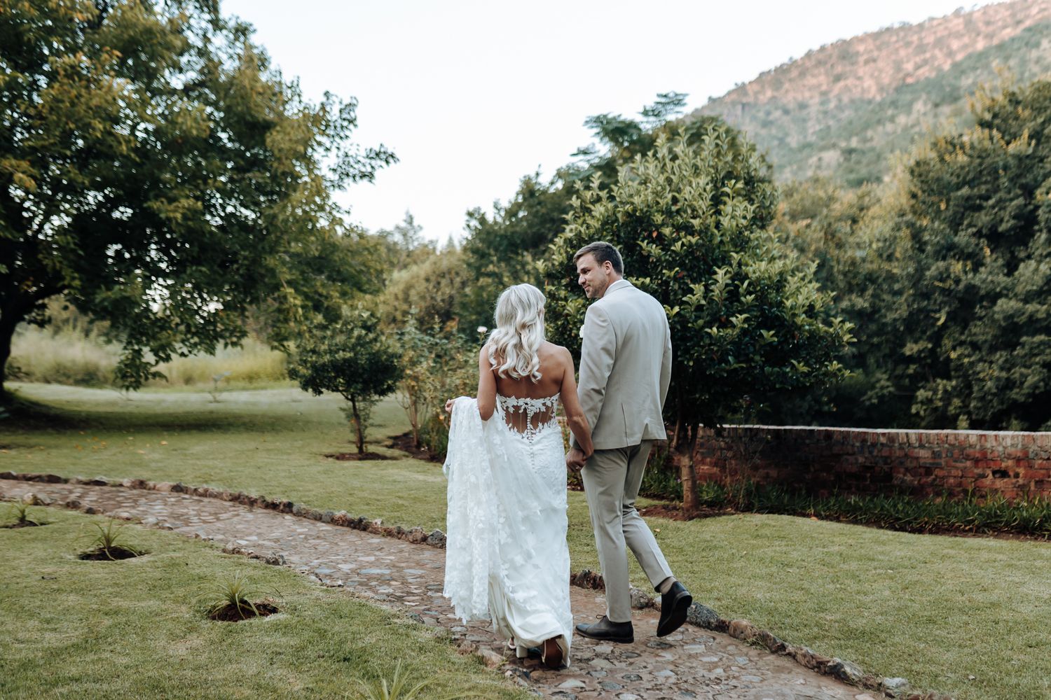 Couple strolling down rustic garden pathway surrounded by lush greenery and mountains.