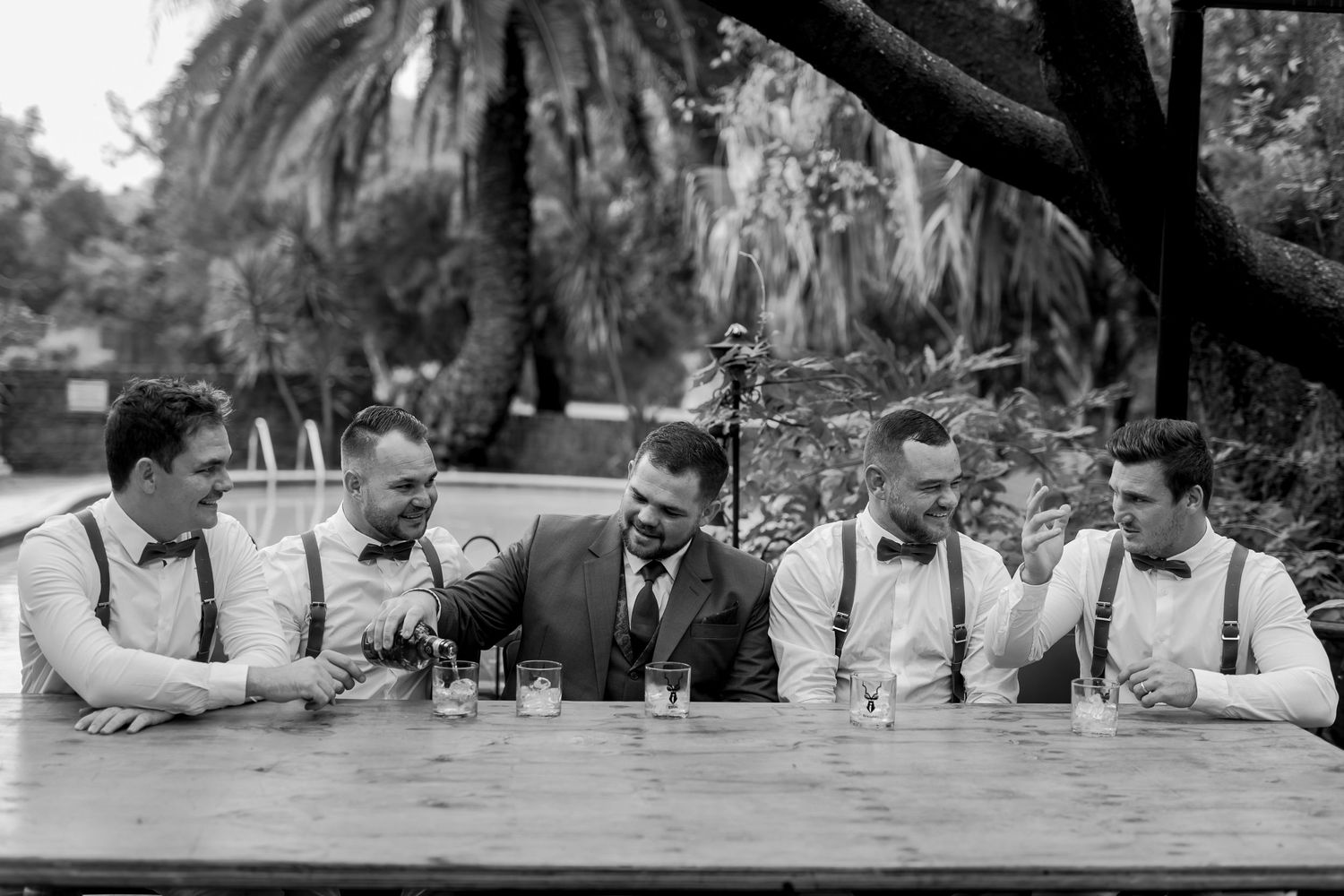 Black and white photo of groomsmen sharing drinks at an outdoor table under palm trees.