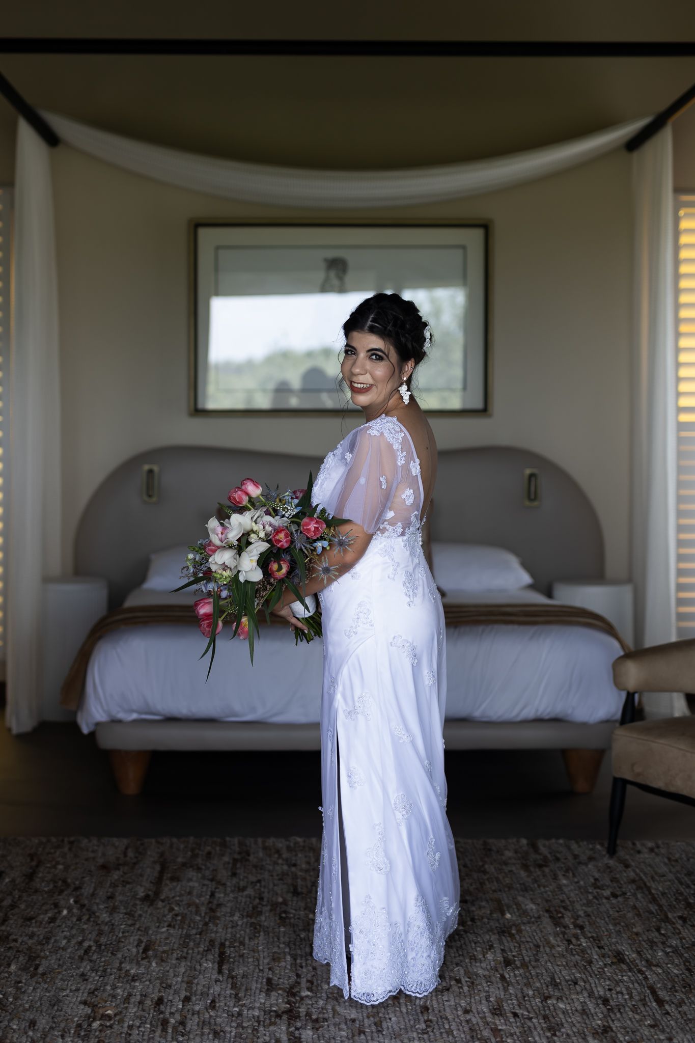 A bride poses in an elegant white lace gown with sheer details in front of a bed holding a floral bouquet.