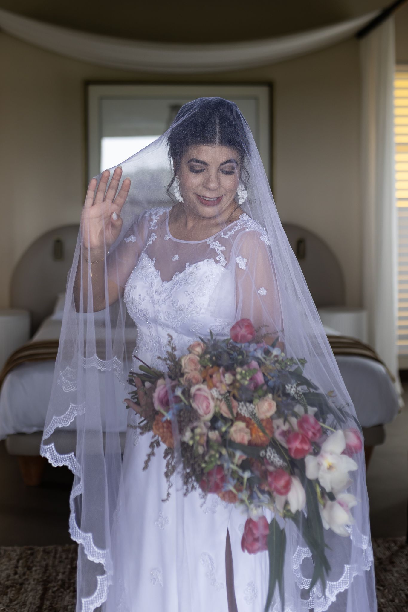 Bride in lace white dress and veil holds a lush bouquet of pink and orange flowers.