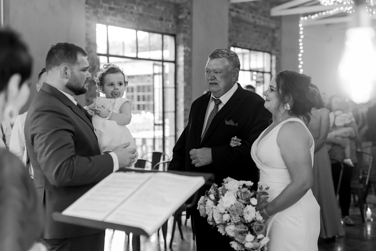 A black and white wedding ceremony moment shows a couple and baby during intimate exchange.