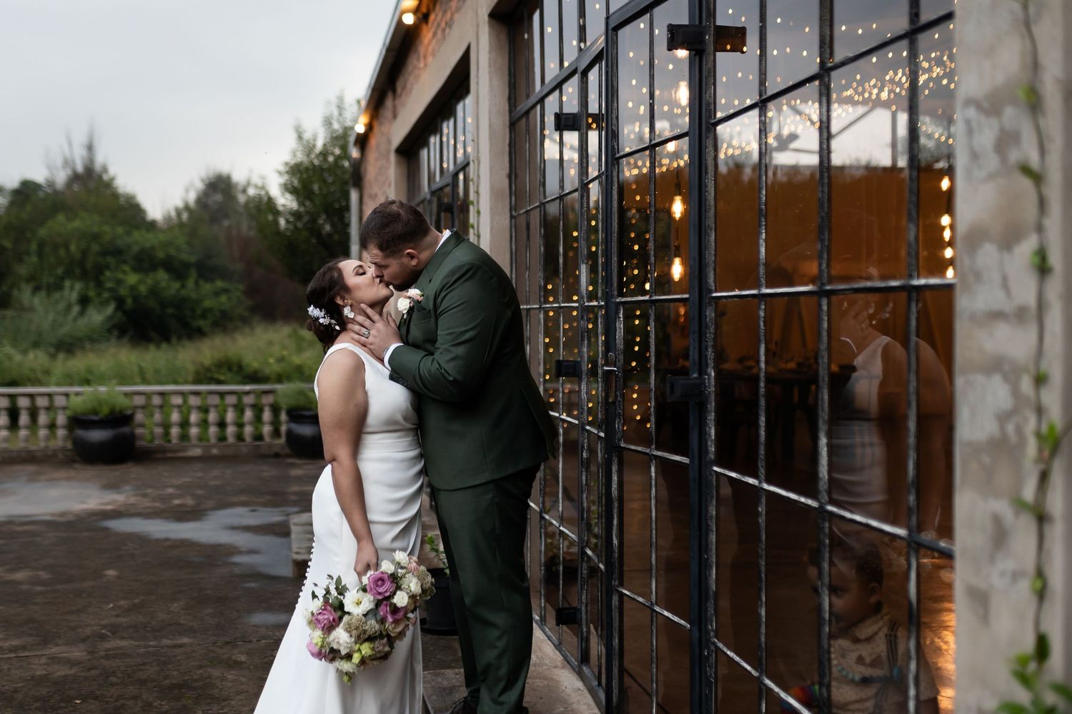 Couple embraces outside wedding venue with string lights glowing through industrial windows.