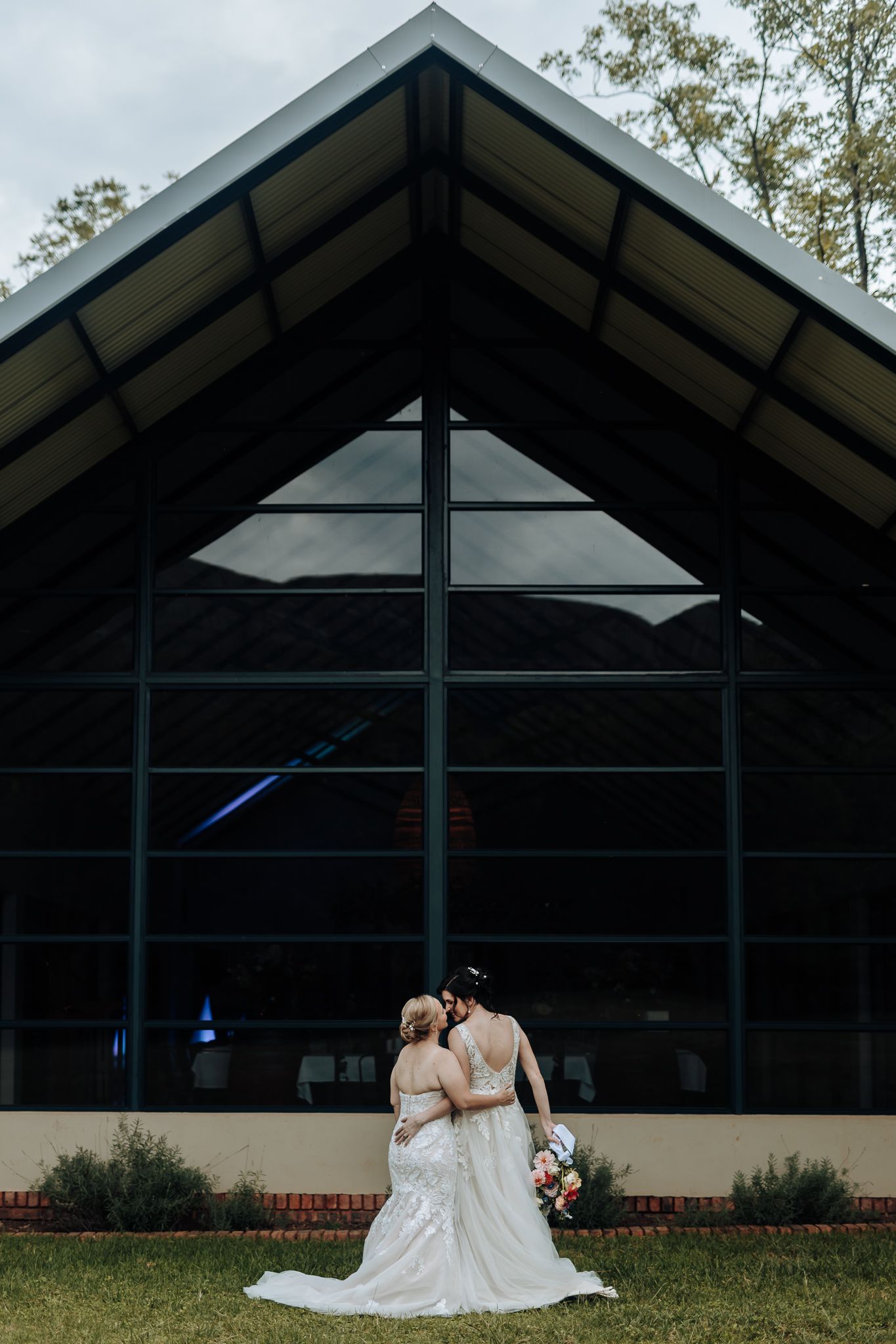 Romantic wedding couple poses against geometric glass building exterior at dusk.