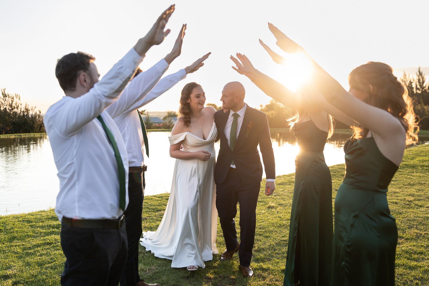 Celebratory moment with wedding party raising arms against sunset by the water.