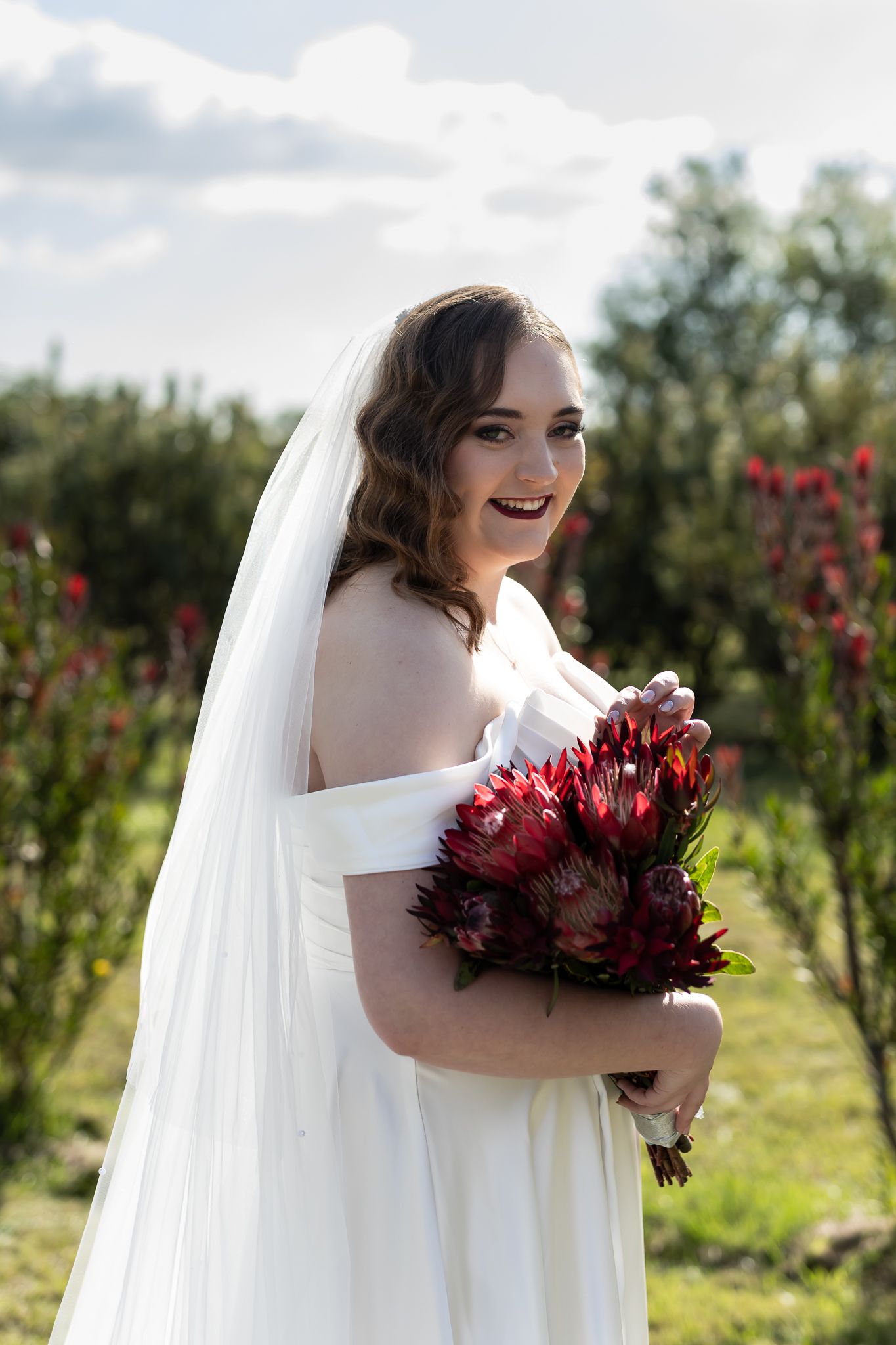 Bride holding burgundy bouquet smiles in white off-shoulder gown among flowering bushes.