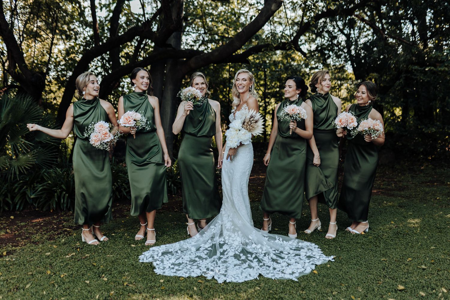 A bridal party in emerald green dresses poses outdoors with the bride's stunning lace train displayed on grass.