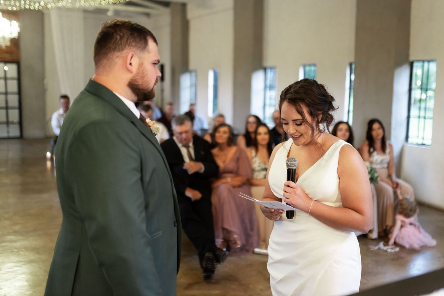 Wedding couple exchanging vows in bright venue with string lights and chandelier.
