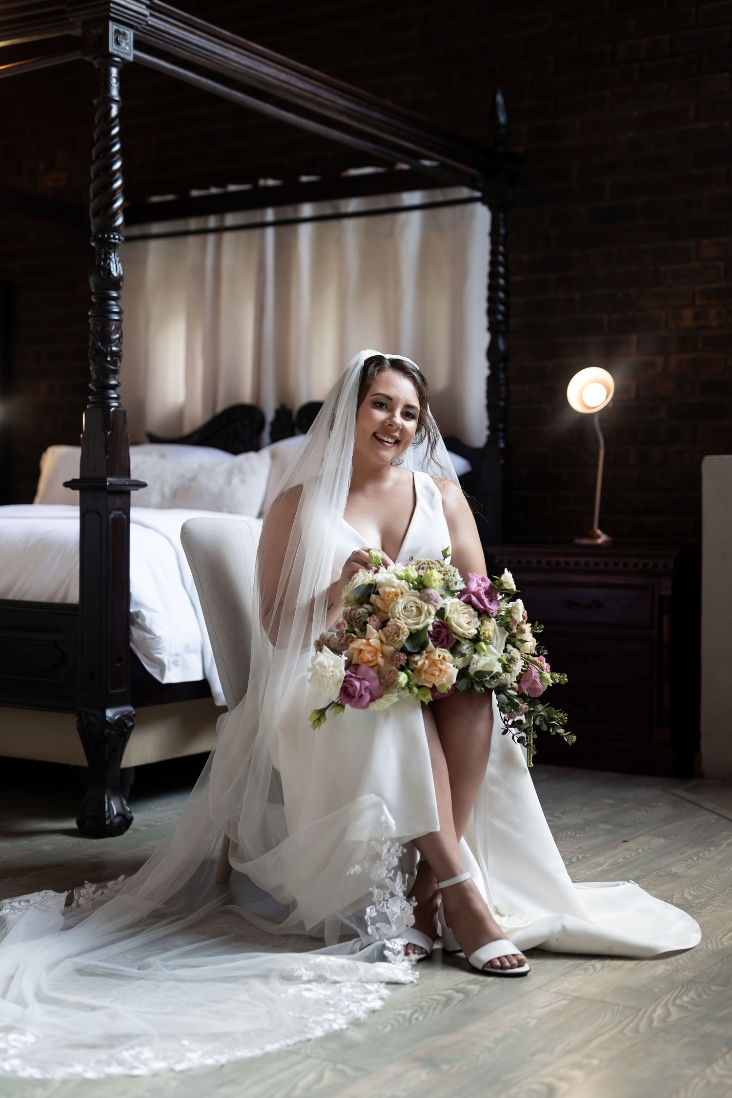 A bride in a white gown and veil sits on a chair holding a bouquet in a room with a four-poster bed.