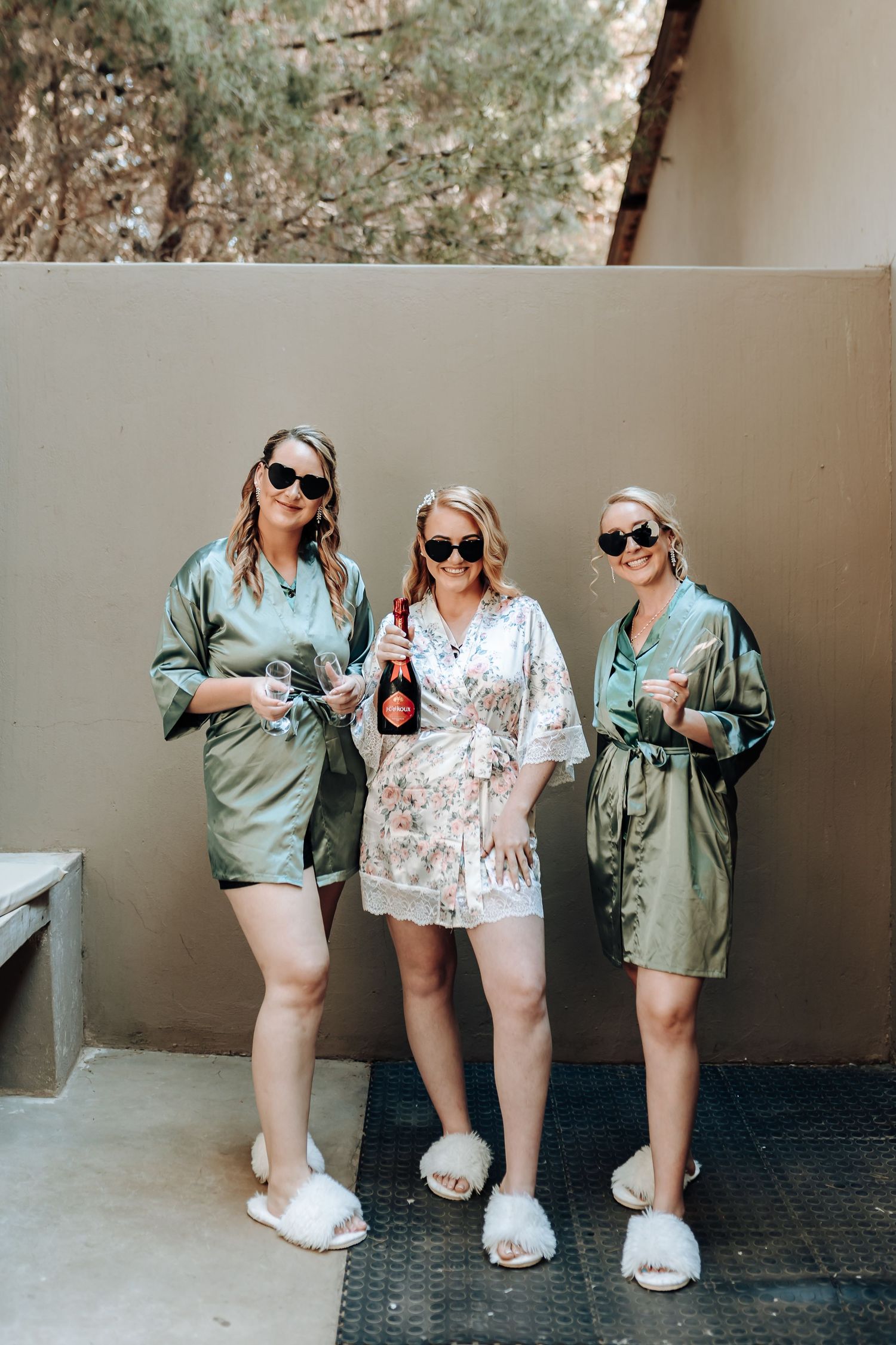 Three friends in matching satin robes and sunglasses pose together while holding drinks outdoors.