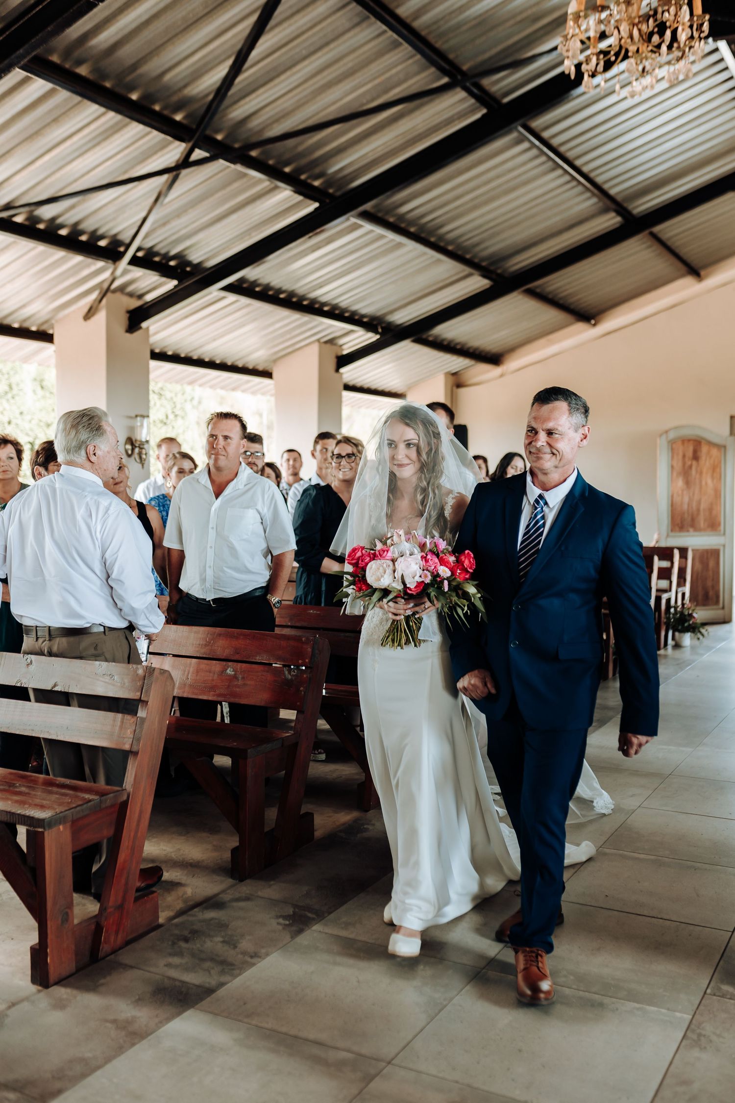 Wedding procession walking down wooden pew-lined aisle in covered venue with chandelier overhead.