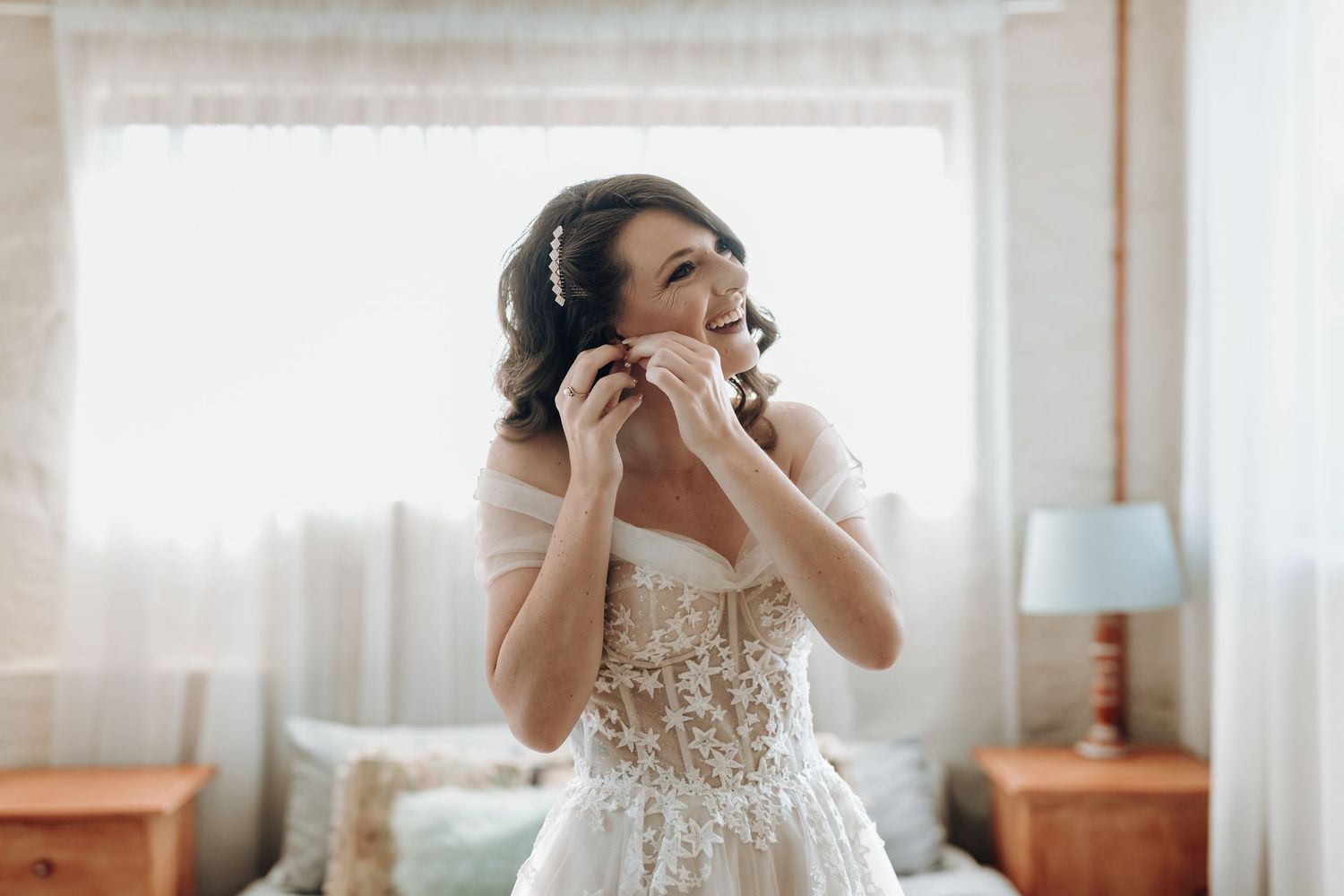 A bridal preparation moment showing a white lace wedding dress and delicate accessories in a sun-lit bedroom.