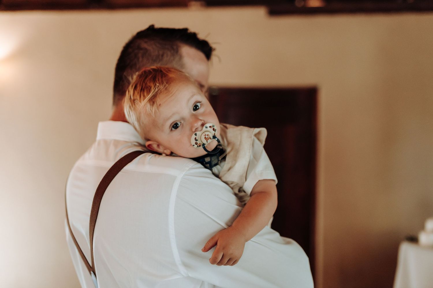A small child with a pacifier rests on someone's shoulder wearing white at a formal event.