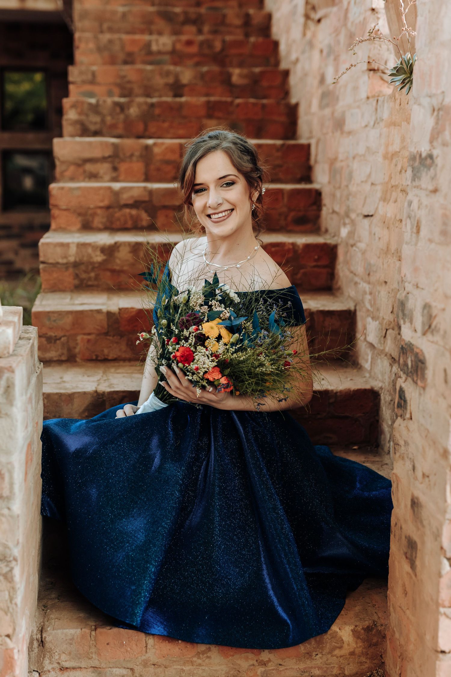Bridesmaid in navy gown sits on brick stairs holding vibrant floral bouquet.