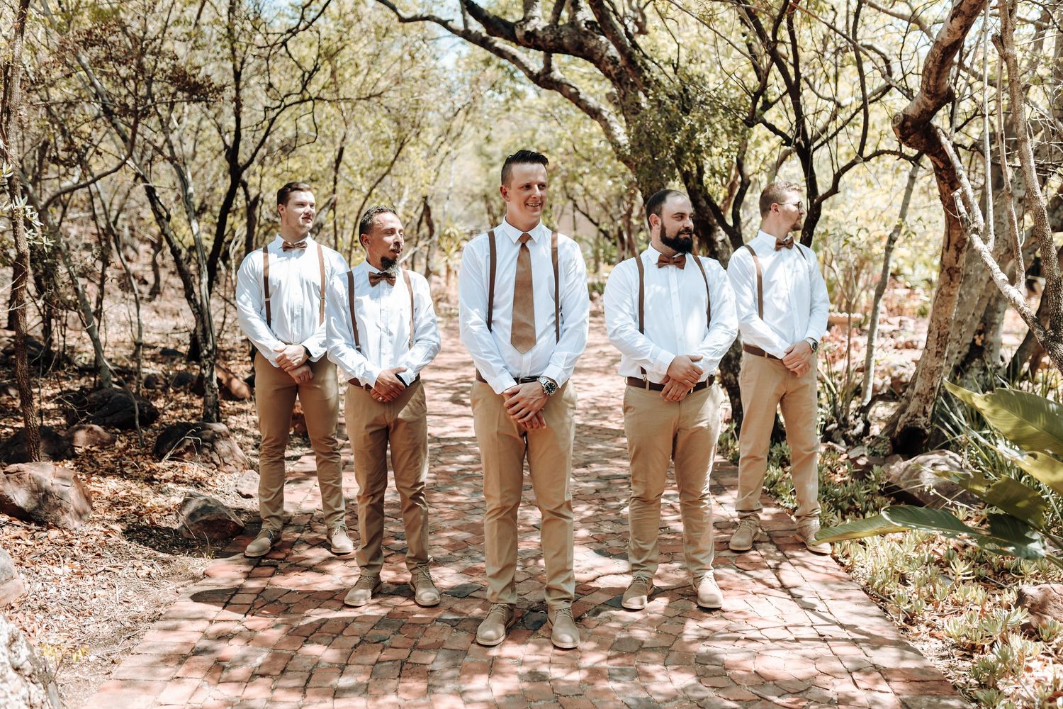 Wedding party in coordinated tan and white outfits pose on a rustic trail.