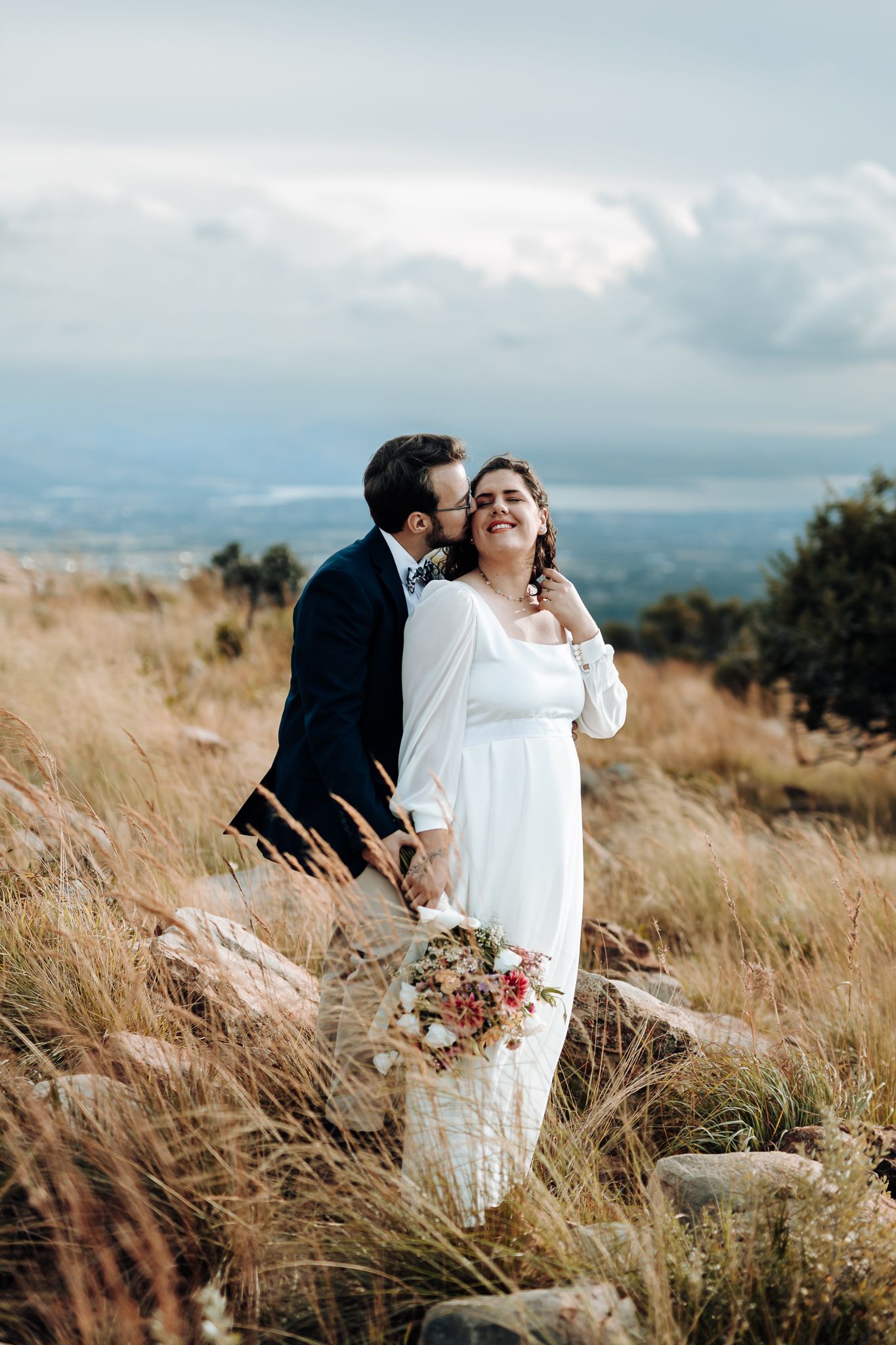 A tender wedding moment captured on a hilltop with dramatic cloudy skies and mountain views.