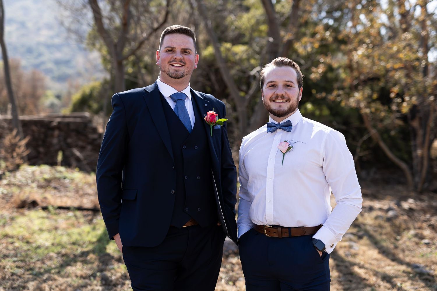 A pair in formal suits standing outside on a path with autumn trees and foliage in the background.