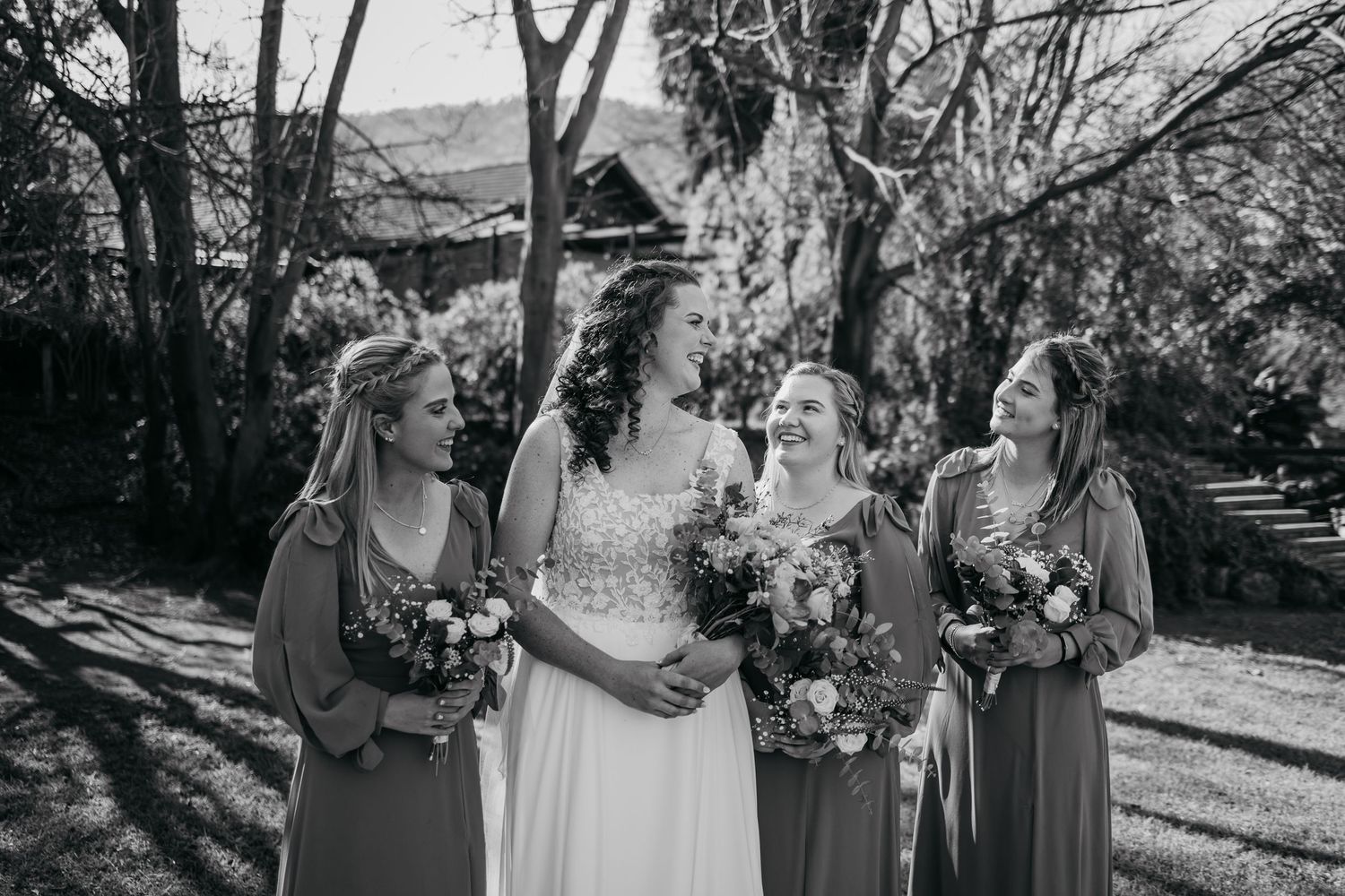 A black and white photo of a bride and bridesmaids holding bouquets while sharing a joyful moment outdoors.