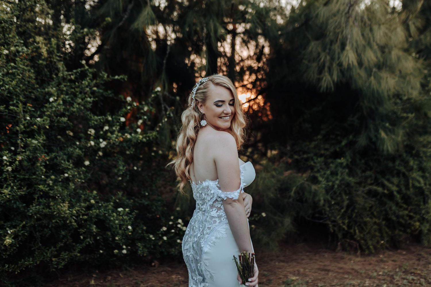 Bride in a fitted lace gown poses elegantly against a sunset garden backdrop.