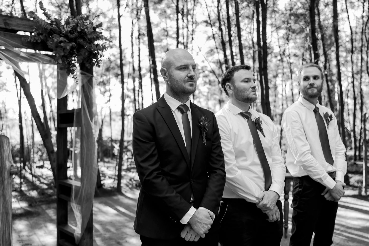 Three groomsmen in formal attire pose together in a sunlit forest setting.