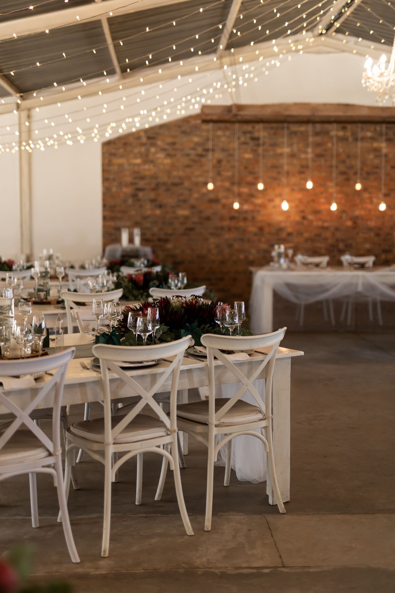 Wedding reception space with white cross-back chairs, fairy lights, and brick accent wall.